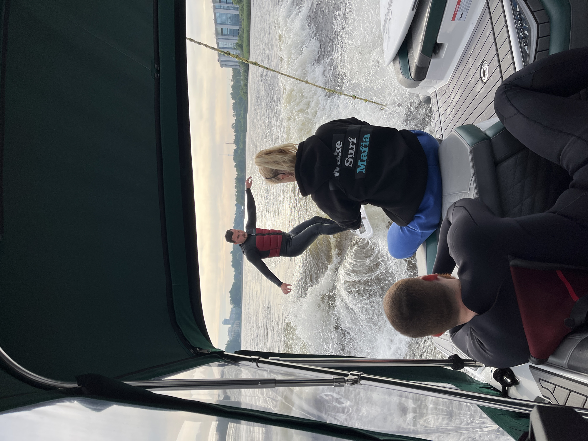 In Saint Petersburg, Russia, on a waterway, a man is wake surfing behind a boat. He is wearing a black wetsuit with a red life vest. In the foreground, a woman with blonde hair in a ponytail, wearing a black hoodie with "Wake Surf Mafia" printed on the back, sits on the edge of the boat. Two other individuals in wetsuits are seated further back in the boat. The boat has a dark green canopy. Buildings and trees line the distant shore. The sky is overcast.