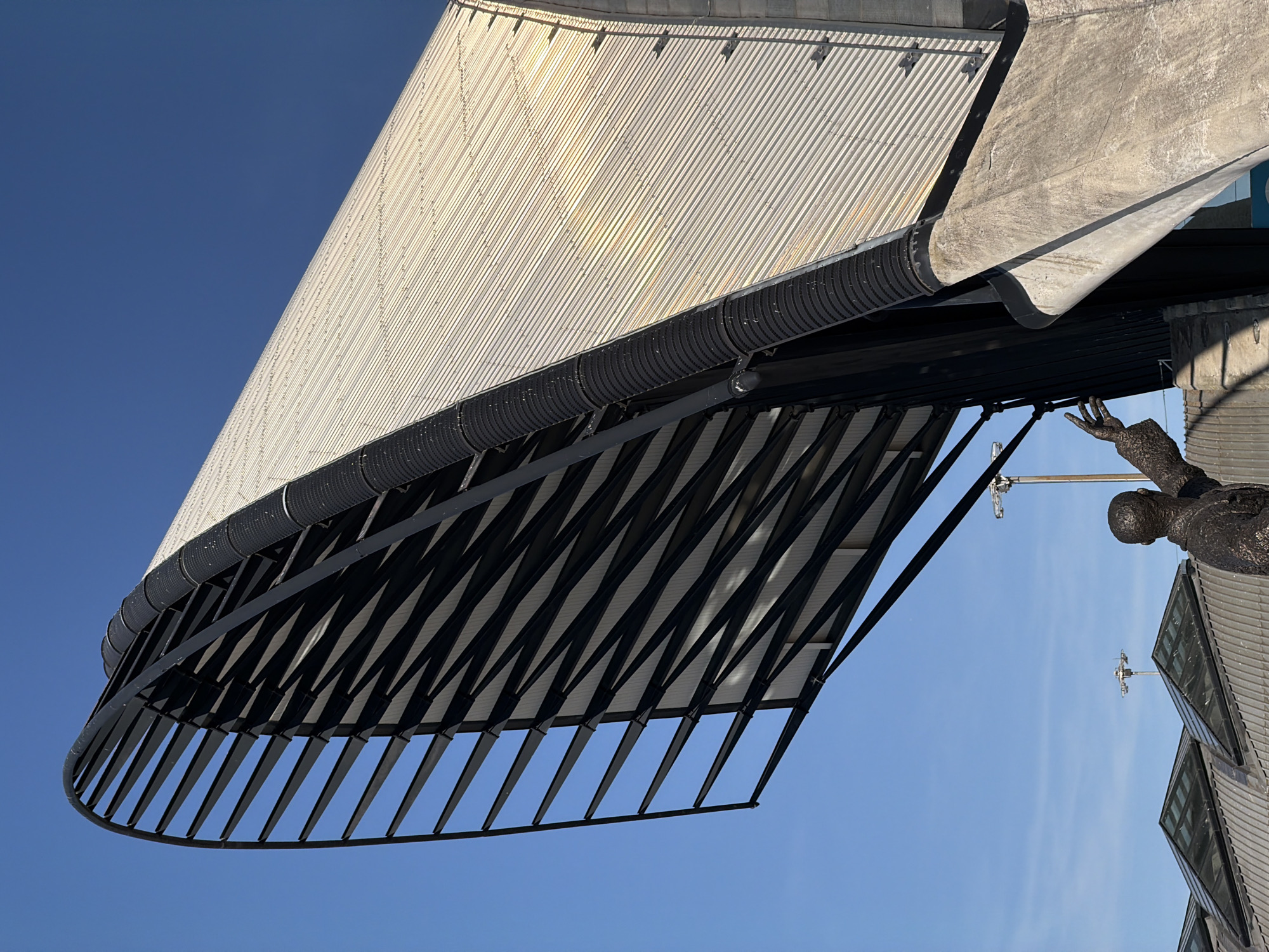 An outdoor, low-angle, eye-level shot captures the side and roof of a modern, curved building against a clear blue sky in Colombier-Saugnieu, France. The building features a silver, corrugated metal roof and a black, ribbed vertical pipe running along its side. To the left of the building, a dark, skeletal structure made of metal beams forms an arched opening. In the foreground, a bronze statue of a man with outstretched arms is visible, with parts of other buildings and skylights in the background. Two lampposts are also visible in the distance. The lighting suggests it is daytime.