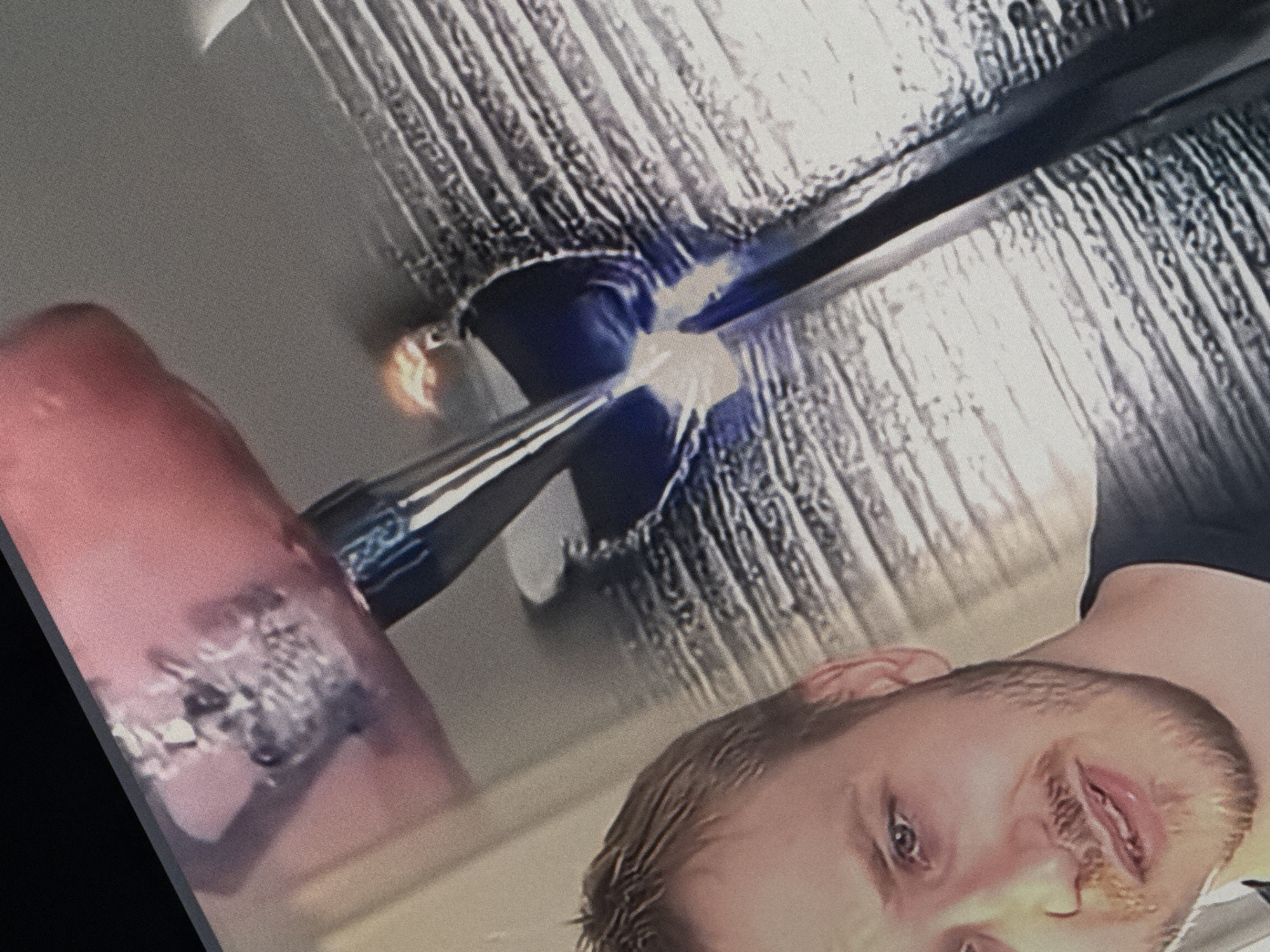 The image shows a close-up of a person's face and a metal object with a blue light. The person is a white male with light brown hair and a beard, wearing a dark t-shirt. He is looking towards the camera with his mouth slightly open. The metal object appears to be a tool or weapon, with a reflective surface and a glowing blue light at its tip. The background is a textured surface, possibly a wall or a piece of equipment. There is no identifiable information in the image that would indicate its location in Saratov, Russia, or any other specific city.