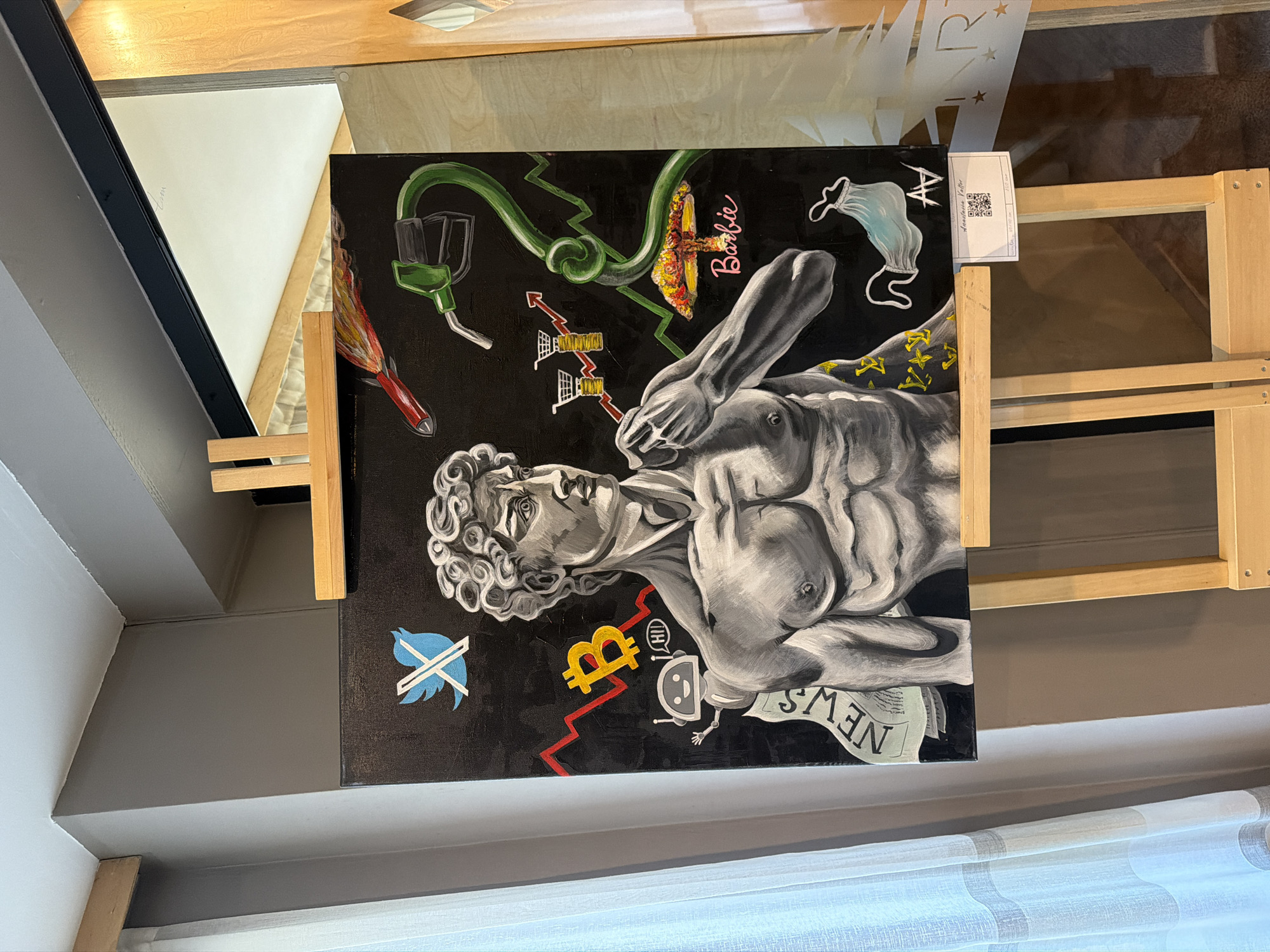 The image shows an artwork displayed on an easel. The artwork features a monochrome rendering of Michelangelo's David, superimposed on a black background adorned with contemporary symbols. These symbols include a stylized Twitter logo, a Bitcoin symbol, a robot figure, a medical mask, a gas pump nozzle, and a Barbie logo. A newspaper with the word "NEWS" is also integrated into the artwork. The easel is made of light-colored wood. The artwork is placed in a setting with a gray wall and a window covered with white curtains in the bottom portion of the frame. The location is described as Unknown, Unknown.
