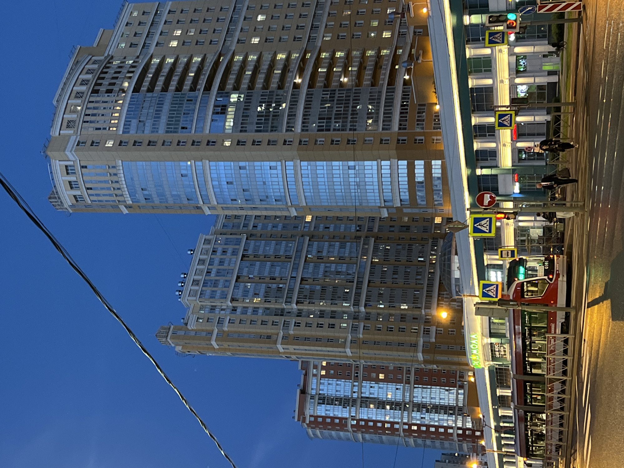 This image shows a street scene at dusk with modern apartment buildings and a tram. The buildings are tall and feature many windows, some of which are illuminated, suggesting it is evening. The sky is a deep blue, indicating twilight.
In the foreground, a tram with red and white accents is stopped at a station. People can be seen inside and near the tram. Several street signs are visible, including traffic lights, pedestrian crossing signs, and a red circular sign with a diagonal line through it, commonly indicating a prohibition.
The buildings have a contemporary architectural style, with curved facades and extensive use of glass. One building in the background appears to have a dome-like structure on top. A wire runs diagonally across the lower left portion of the image, obscuring part of the sky and buildings.
The overall scene suggests an urban environment in Russia, likely a residential area with public transportation. The lighting and the blue of the sky point to the time of day being around sunset or just after. The presence of people and the tram suggests daily life is ongoing. There is some visible text on the tram and building facades, though it is not clearly legible.