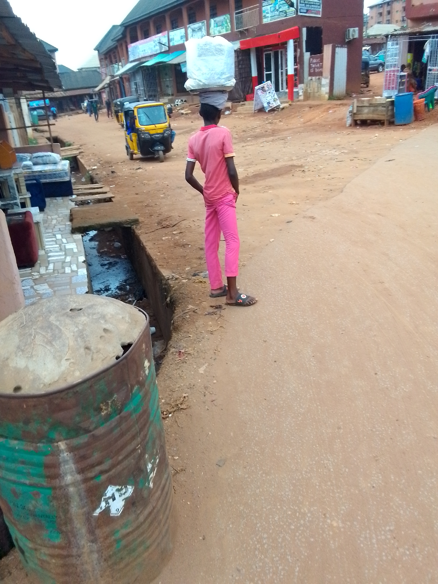 The image depicts an active street scene in Onitsha, Nigeria, characterized by an unpaved, reddish-brown dirt road. In the immediate foreground, a person, seen from behind, wears a pink t-shirt and pink trousers, carrying a large, white, bundled package on their head. They are wearing dark sandals. To their left, a heavily rusted, partially green and brown metal barrel sits beside an open concrete drainage ditch filled with murky water.
Further down the road, a yellow and black tricycle, commonly known as a Keke Napep, is visible, suggesting traffic on the street. The buildings lining the road are predominantly multi-story commercial structures, with shops and businesses at ground level. Some storefronts display goods, such as mattresses and household items, outside. The architecture includes brown and red facades, with one shop featuring a red and white entryway.
The time of day appears to be daytime, with natural, somewhat diffused light, indicating either an overcast sky or a time other than harsh midday sun. The scene reflects a typical urban or semi-urban environment in Nigeria, with the dusty road and style of transport and commerce acting as key location cues.
Visible text includes a prominent sign on a building in the mid-ground that reads "PUBLIC TOILET IS HERE," with additional, less clear text below it. Further down the street, a partially obscured sign on a blue structure appears to read "PUBLIC MARKET." A white sticker with an indistinct logo, possibly stylized letters, is affixed to the side of the barrel in the foreground.