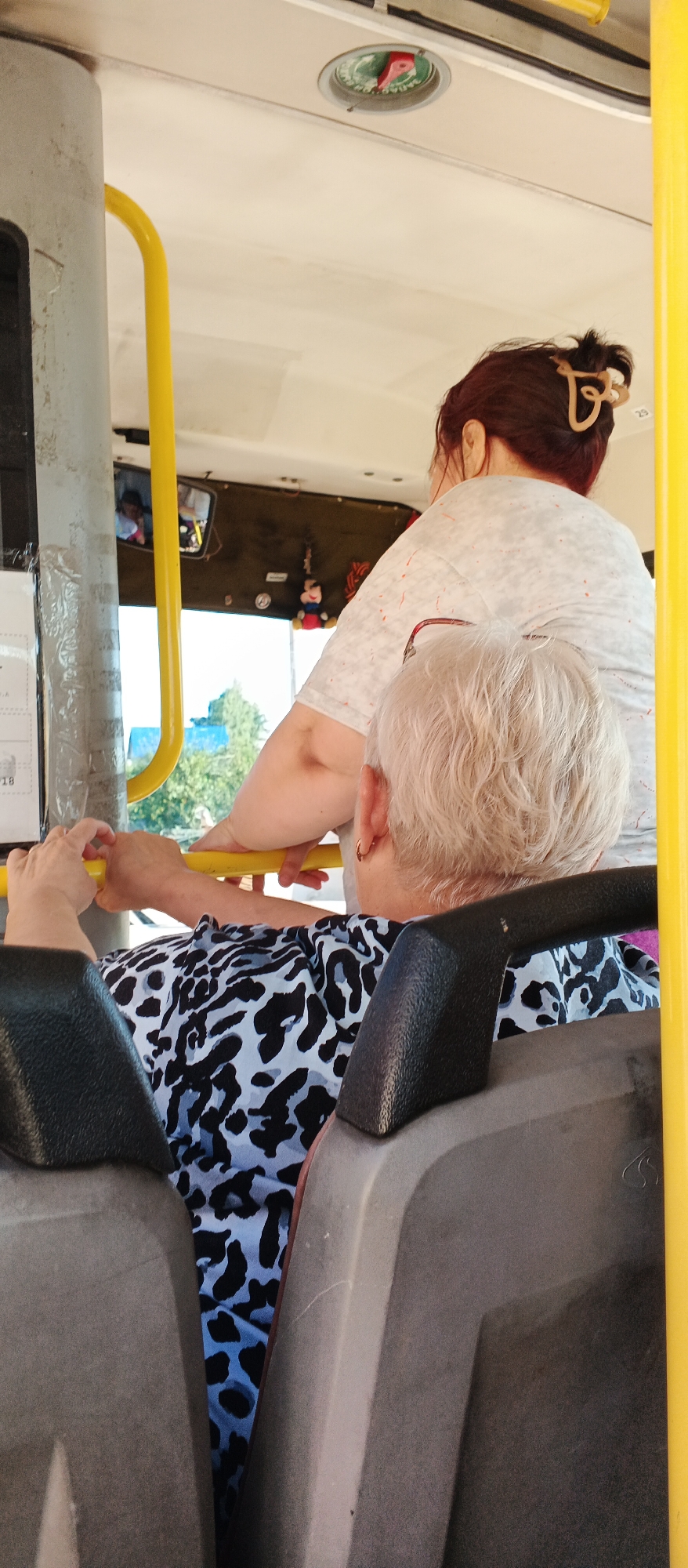 The image shows a view from inside a bus in the city of Kiyasovo, Russia. In the foreground, two seats are visible, one on the left and one on the right, with a gap between them. A person's hands are resting on a yellow grab rail in the left foreground. In the middle ground, an older woman with short, light-colored hair is seated and facing away from the camera. She is wearing a black and blue leopard print top. Behind her, another person with dark hair is standing and also facing away from the camera. This person is wearing a light-colored t-shirt with red speckles. A yellow grab rail is visible running horizontally across the middle of the image. In the background, a mirror reflects a window and some trees. A small Mickey Mouse toy is hanging from an object in the background. The bus interior appears to be well-lit.