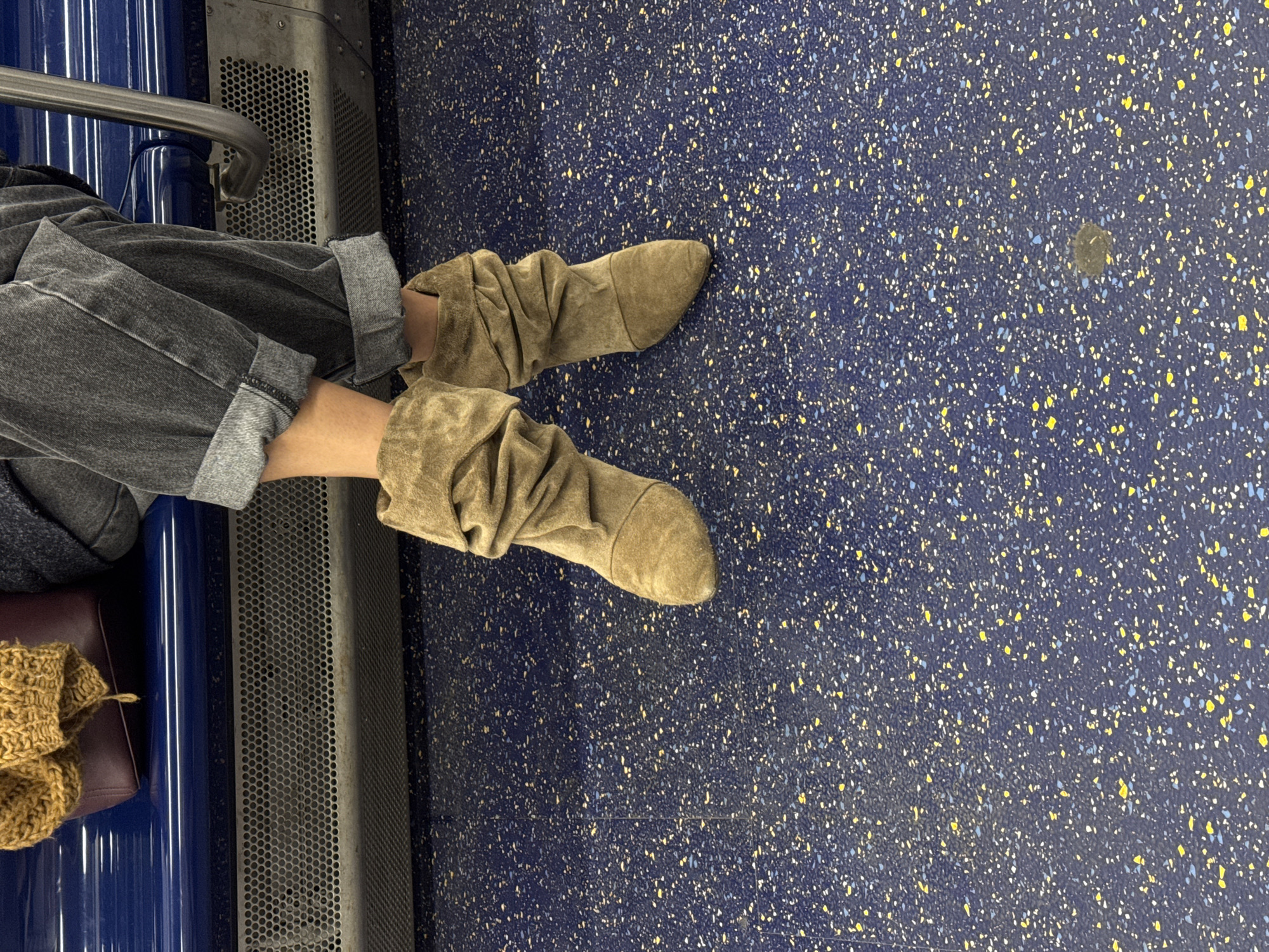 The image shows a person seated on what appears to be public transportation, possibly a subway or train. The person is wearing cuffed gray jeans and slouchy tan boots. Their feet are extended onto the speckled blue and yellow floor of the vehicle. A metal safety bar and a perforated metal panel are visible to the left of the person. A brown knitted item rests on the seat.
Since the prompt includes the city of Hoboken, United States of America, the image could be from the Port Authority Trans-Hudson (PATH) train that serves the city. However, this cannot be confirmed from the image alone.