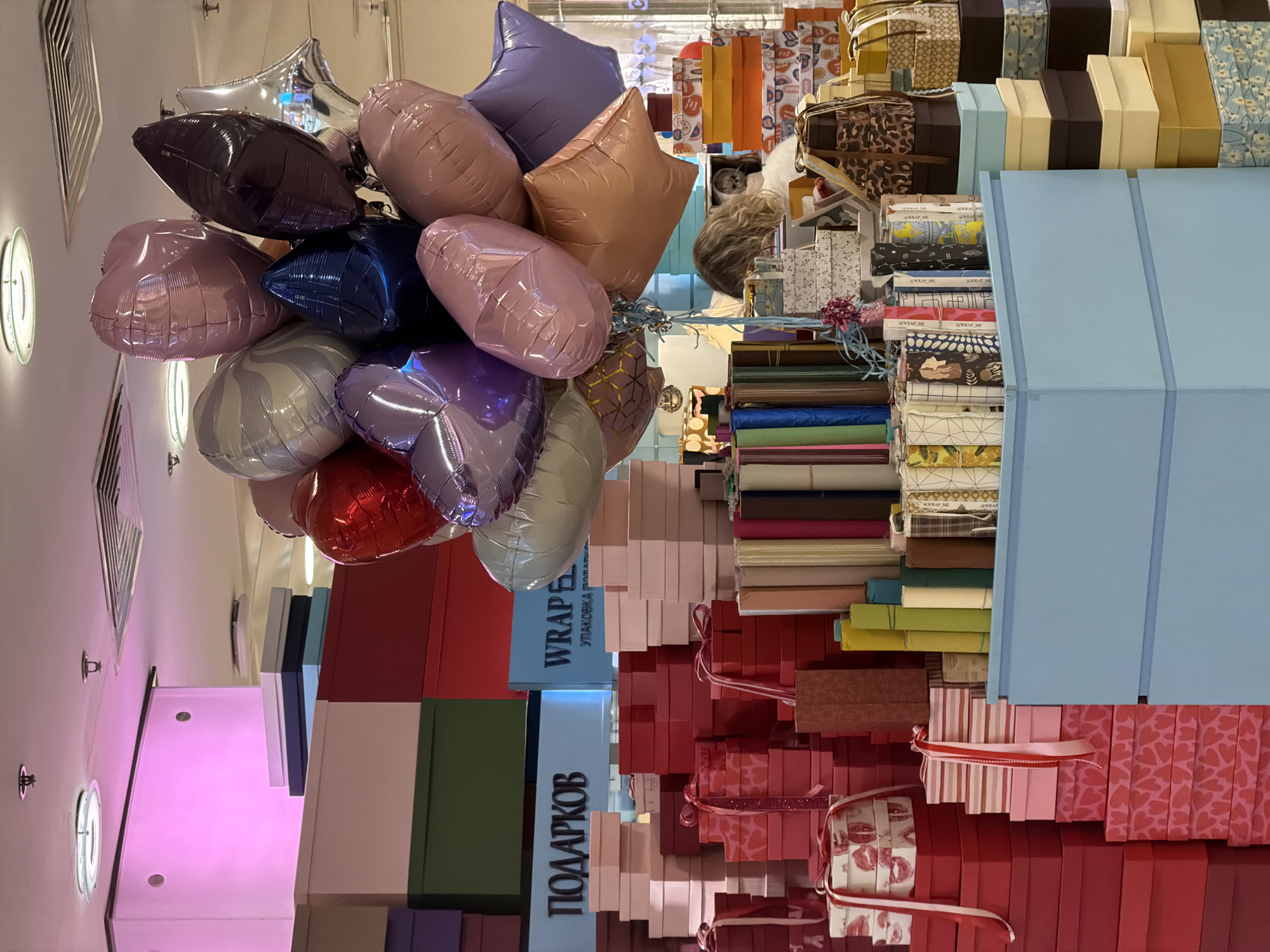 The image shows a scene inside a retail store, likely a gift shop, in Moscow, Russia. The foreground features a cluster of colorful, shiny balloons in various shapes (hearts, stars) and colors (purple, pink, silver, red, blue, gold). The background is filled with neatly stacked boxes of different sizes and colors (red, green, pink, white, blue). Some of the boxes have ribbons and decorative patterns. Between the balloons and the boxes, there are rolls of gift-wrapping paper in different shades. The word "ПОДАРКОВ" (Gifts in Russian) is visible on one of the boxes. There are hanging lights on the ceiling. The overall impression is of a store specializing in gift wrapping and accessories.
