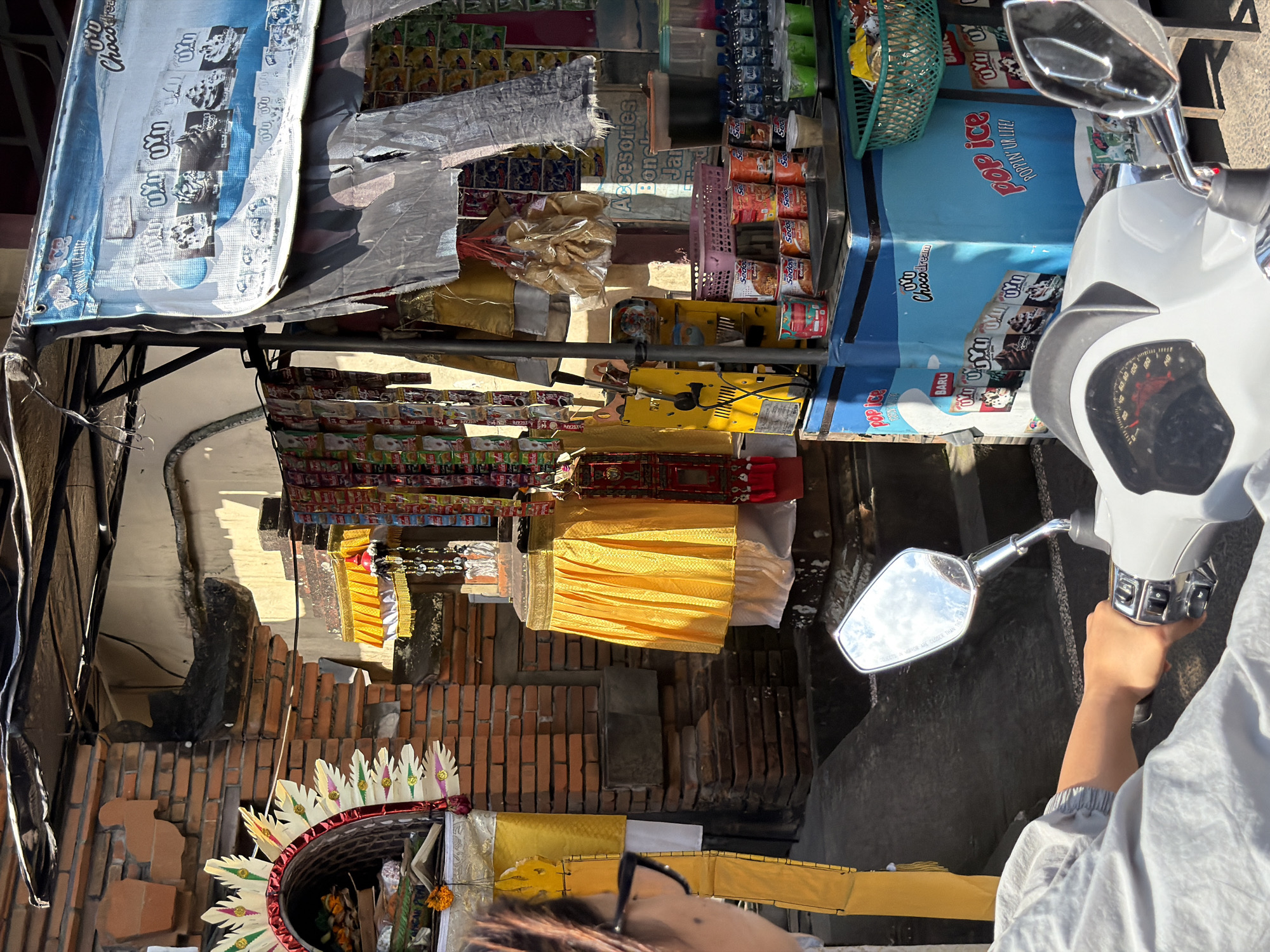 A person wearing glasses and a light grey jacket is riding a white scooter and looking to the left. The scooter's handlebar and part of its dashboard are visible. The person is near a street stall in Ubud, Indonesia. The stall displays various products, including packaged food items, drinks in bottles and cups, and a blue freezer with ice cream packaging. Behind the stall, a brick building is visible with decorative elements on its facade. The scene is brightly lit by sunlight, casting shadows on the ground.
