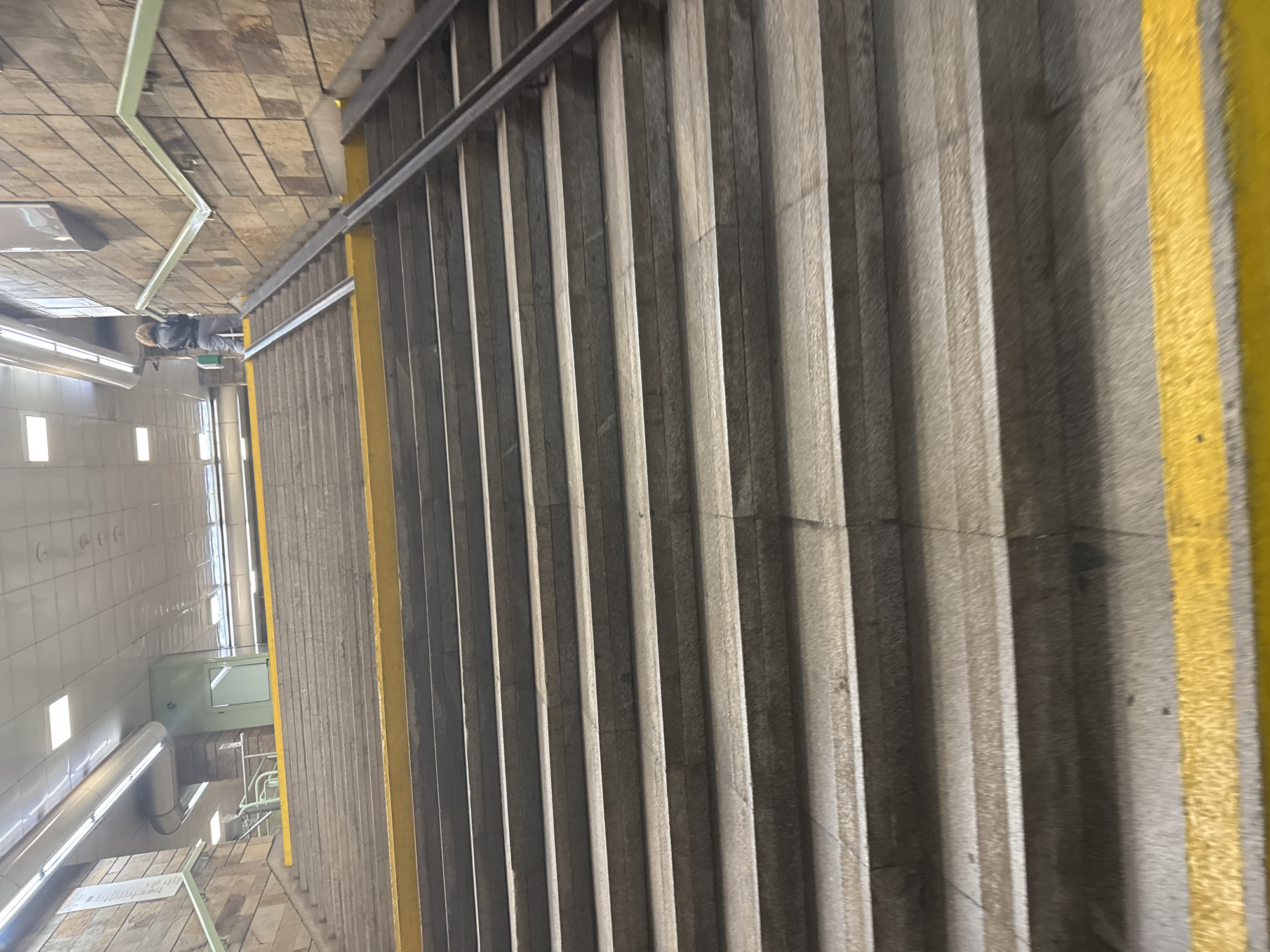 The image depicts a staircase, likely in an underground or subway station in Sofia, Bulgaria. The staircase has a metal handrail and a yellow safety stripe along the edge of the steps. The walls are tiled in white, and there are visible pipes and electrical boxes. At the top of the stairs, a person wearing a gray jacket or uniform is visible.