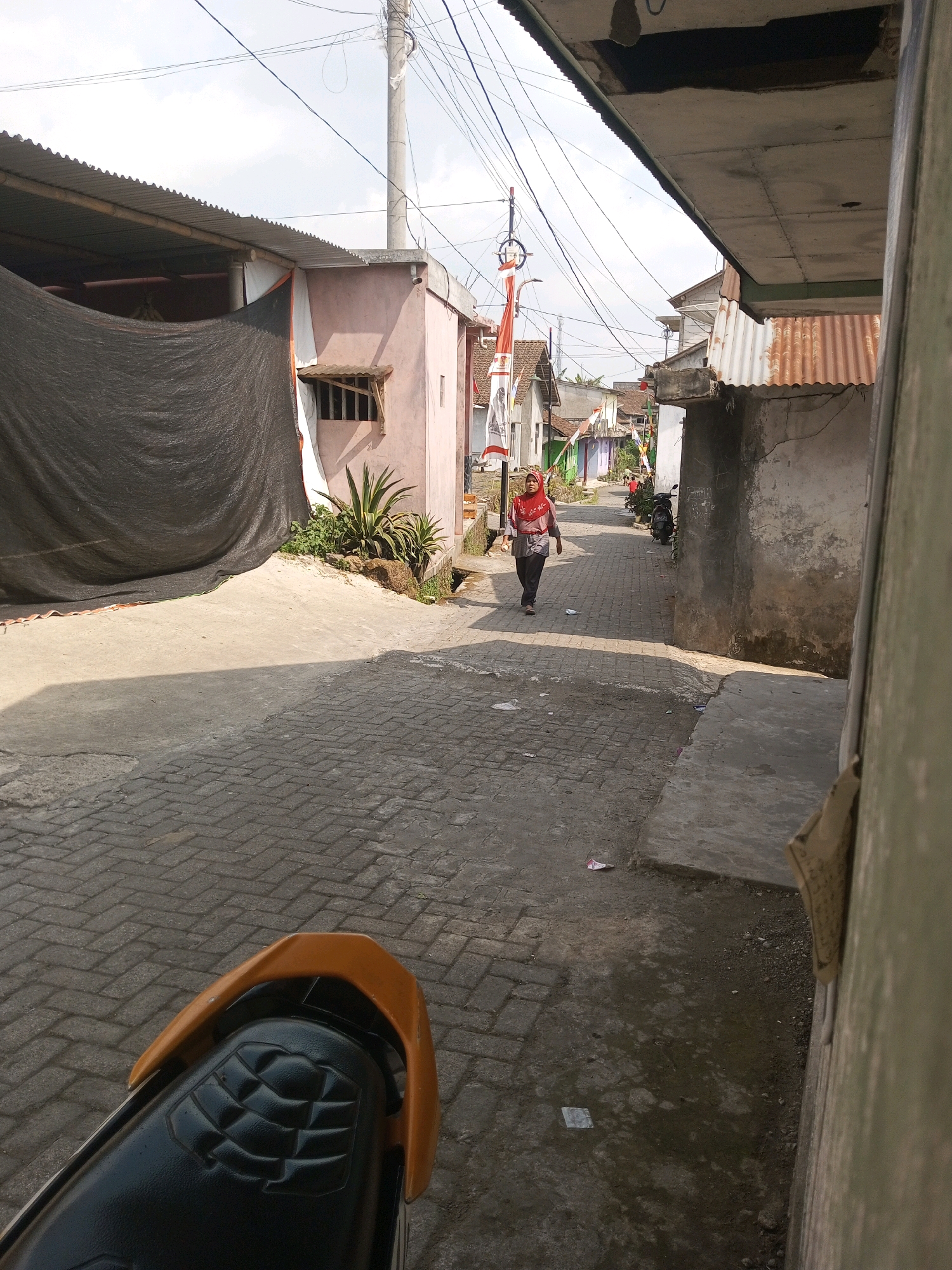 The image depicts a narrow, paved alleyway in Andongsili, Indonesia. A woman wearing a red hijab and a patterned top walks away from the viewer down the alley. She is on the left side of the street, with a pink building to her left and a gray, weathered building to her right. The alley is lined with small residential structures, some with corrugated metal roofs. Indonesian flags are visible, attached to poles along the street. A motorcycle is parked on the right side of the alley. In the foreground, the rear seat and part of the frame of a yellow motorcycle are visible, suggesting the photo was taken from a stationary motorcycle. The ground is paved with interlocking bricks and some areas of dirt. The sky is clear with scattered clouds, and the sun casts shadows across the scene.