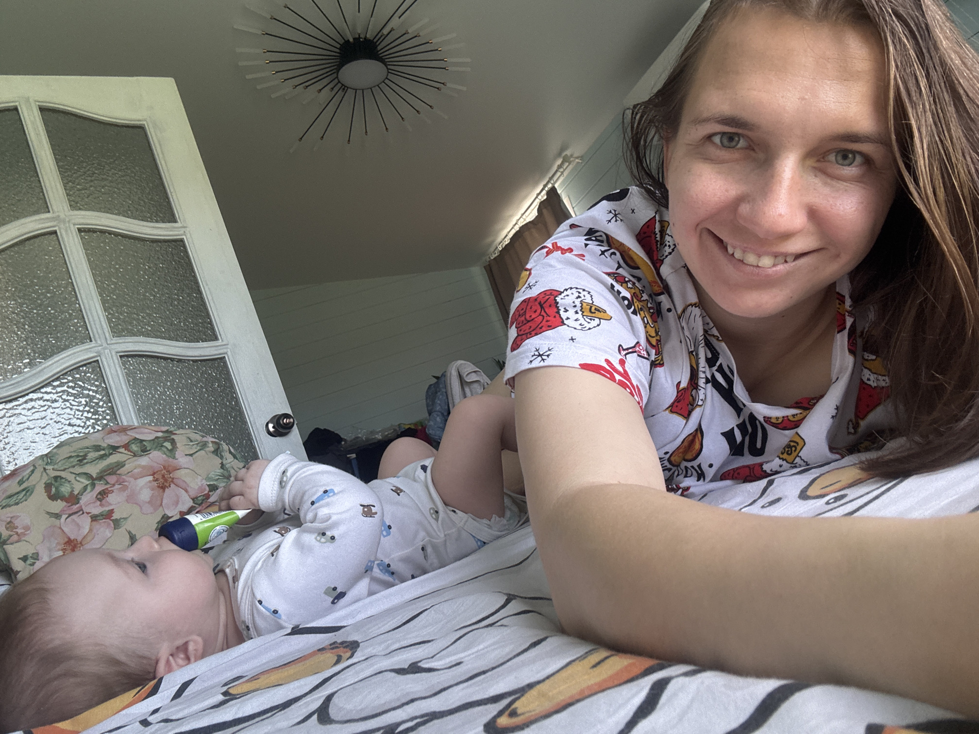 This image captures a woman taking a selfie with a baby in an indoor setting, likely a home in Раздолье, Russia. The woman, in the foreground on the right, smiles warmly at the camera. She has long brown hair and light-colored eyes and wears a white short-sleeved top with festive patterns, including Santa Claus figures and snowflakes, with some text possibly including "SANTA'S BEST".
To her left, a baby lies on a bed, looking towards the left side of the frame, holding a green and blue cylindrical object near its mouth. The baby is dressed in a white long-sleeved onesie with small blue and grey car prints and a white diaper. They are resting on a bed sheet featuring an abstract pattern of black, white, and orange, with a floral pillow visible behind the baby's head.
In the background, a white door with frosted glass panels is partially visible on the left. Above, a modern, sputnik-style ceiling light fixture with a black base and radiating rods is mounted on a light-colored ceiling. The wall behind the bed is paneled horizontally in a light color. The scene is brightly lit, indicating daytime. The overall atmosphere is intimate and serene, depicting a joyful moment.