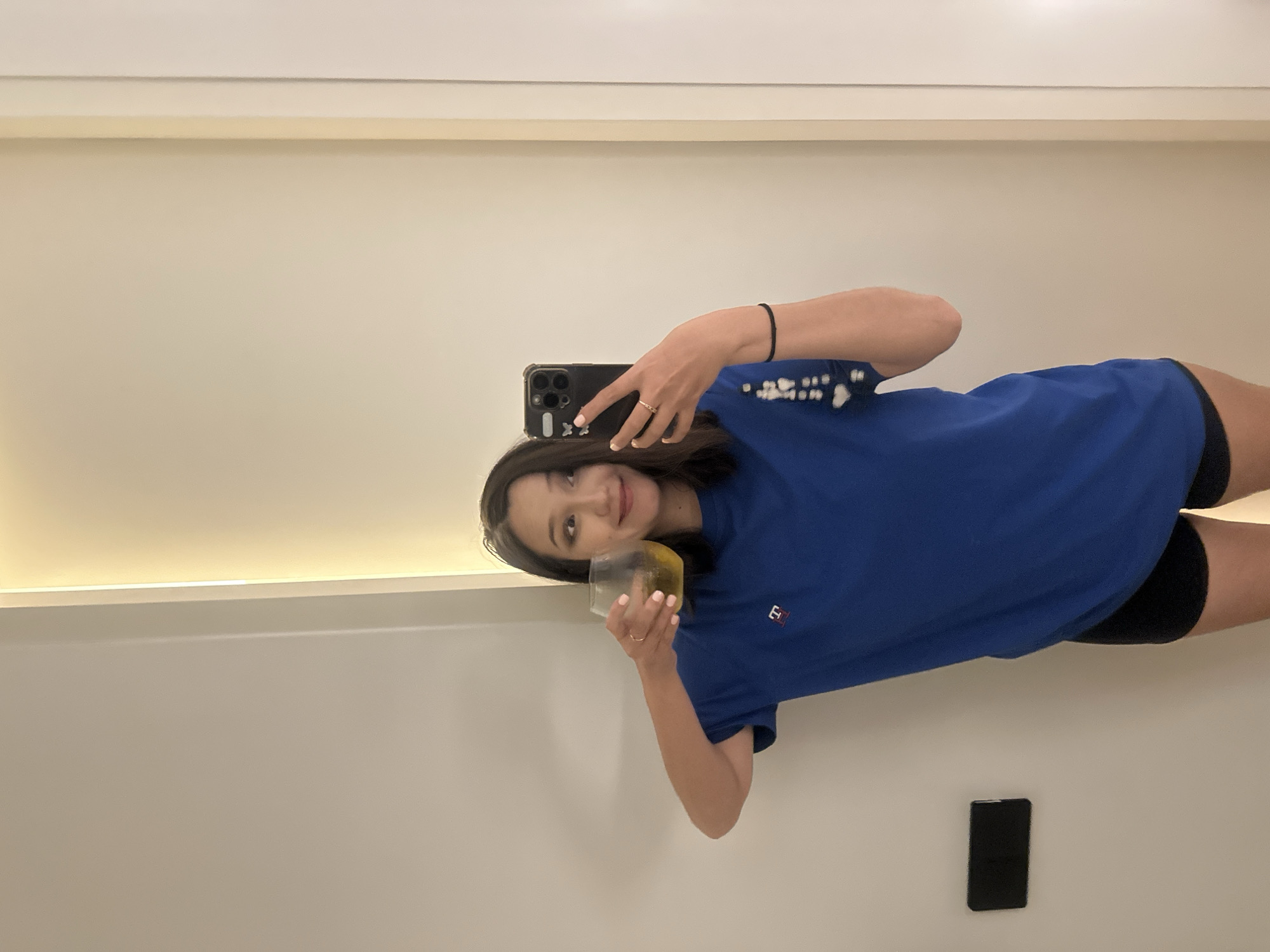 A young woman is taking a selfie in a room. She is wearing a royal blue t-shirt with a white logo on the left chest and black shorts. She is holding a glass of amber-colored liquid in her left hand and her right hand is holding her phone, which is in a black case with white butterfly stickers. She is standing in front of a plain wall with a vertical light fixture to her right. There is a black rectangular object mounted on the wall to her left. The overall environment suggests an indoor setting, possibly a hotel room or apartment. Without further information, the specific location in North Kuta, Indonesia cannot be determined from the image alone.