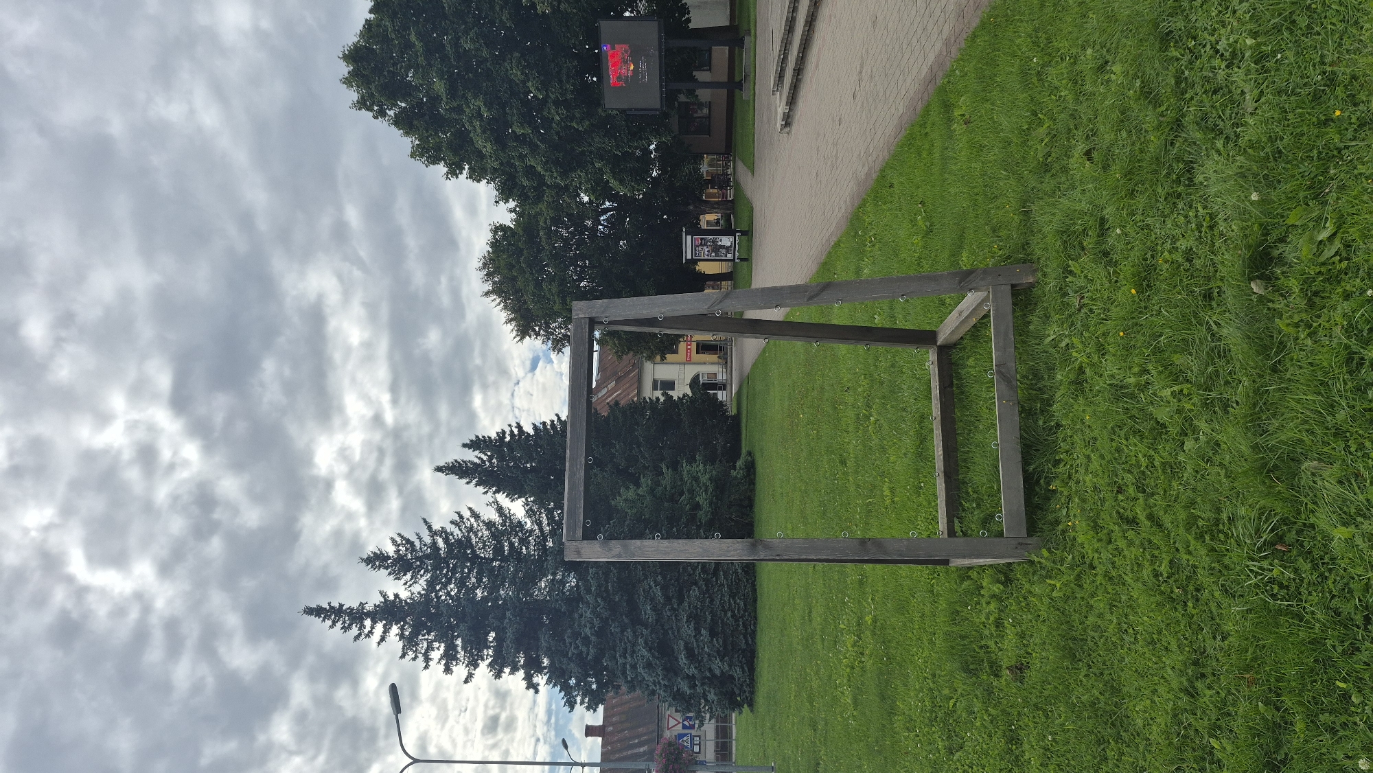 The image captures an outdoor scene in Krāslava, Latvia, likely a public space with a grassy area and a paved walkway. In the foreground, a rustic wooden frame, possibly part of a swing set or an art installation, stands on the lawn. The frame is constructed from thick wooden beams and has metal rings attached to its horizontal supports. Behind the frame, lush green grass covers the ground, with a few scattered yellow wildflowers.
Beyond the grassy area, a paved pathway with interlocking bricks runs diagonally across the scene. To the left of the pathway, several large evergreen trees, possibly spruce or fir, are prominent, with a street lamp visible nearby. Further back, several buildings with red-tiled roofs line a street, suggesting a town or city environment. One building appears to be a commercial establishment with signage.
To the right of the pathway, a large outdoor LED screen is mounted on a pole. The screen displays content, though the details are not clearly discernible. The sky above is overcast with a layer of grey clouds, indicating a cloudy day. There are no people visible in the immediate vicinity of the wooden frame or the surrounding area. The overall impression is of a quiet, public park or plaza in Krāslava.