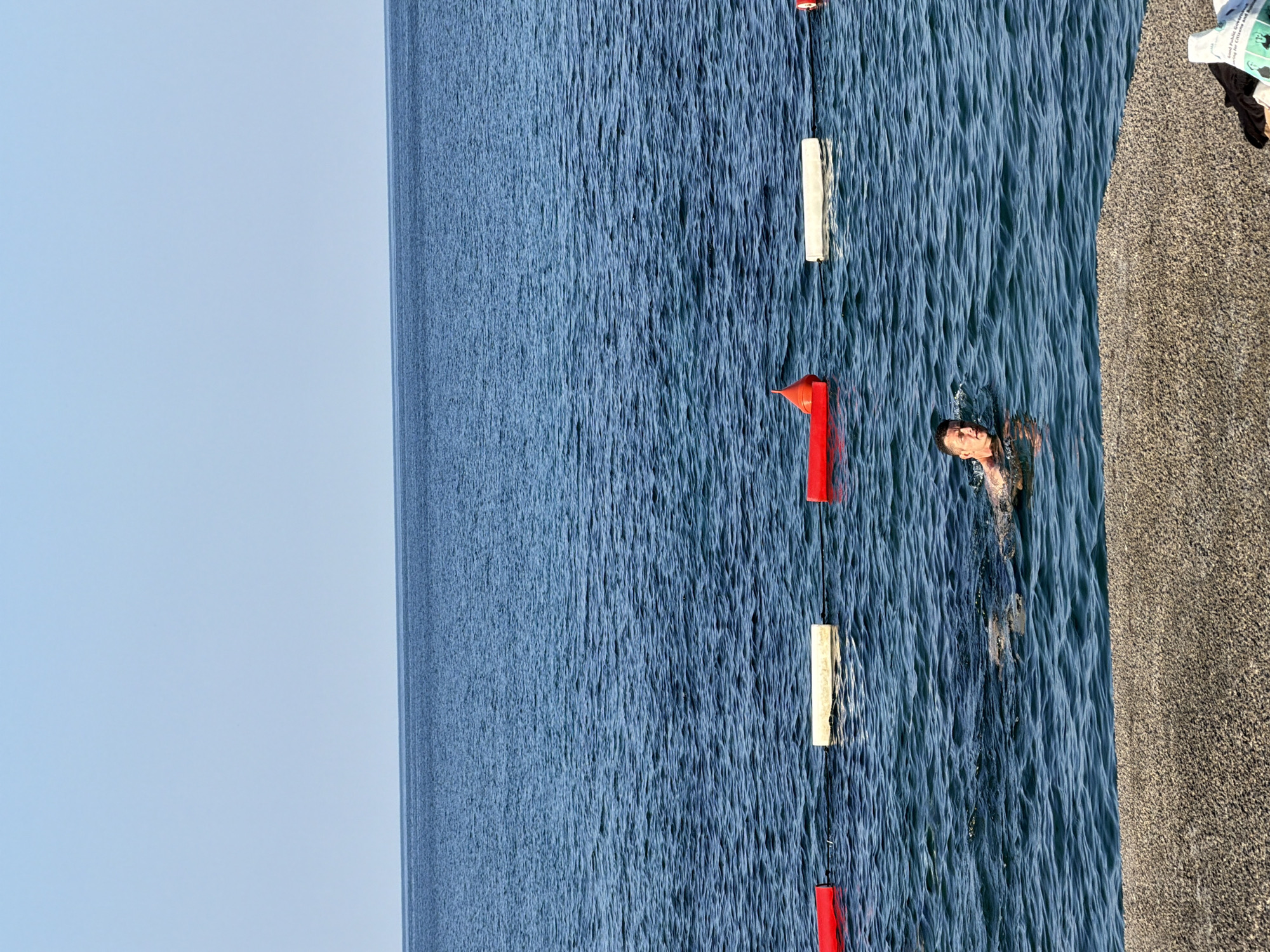 A man swims in the calm, blue waters of the Adriatic Sea near the shoreline of Budva, Montenegro. The man, who appears to be in his late 30s or early 40s, is looking towards the camera with a neutral expression. He is partially submerged in the water, with his head and shoulders visible. In the background, a line of buoys separates a swimming area. The water is clear and the seabed is visible near the shore where a coarse, gravelly embankment is present. The sky above is a clear, pale blue. The overall scene suggests a peaceful, recreational day at the beach.