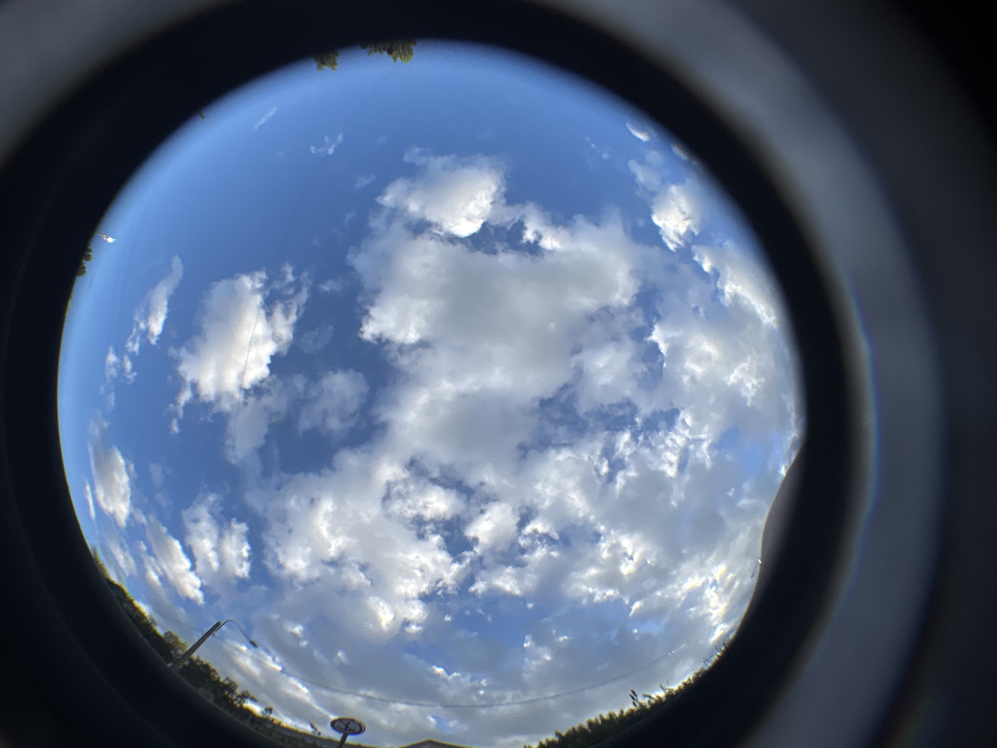 A fisheye lens captures a vast expanse of blue sky dotted with fluffy white clouds, framed by a dark, circular border that suggests the image was taken through a camera lens or a similar optical device. The curvature of the image distorts the horizon, which appears as a dark, blurry line with hints of green foliage and dark vertical structures, possibly trees and utility poles, on the left side. A faint, rainbow-like spectrum of light is visible along the bottom edge of the frame, indicative of lens flare or optical effects. The overall impression is one of a bright, daytime sky with scattered cloud cover. The context of Saint Petersburg, Russia, is not discernible from the visual information provided in the image itself.