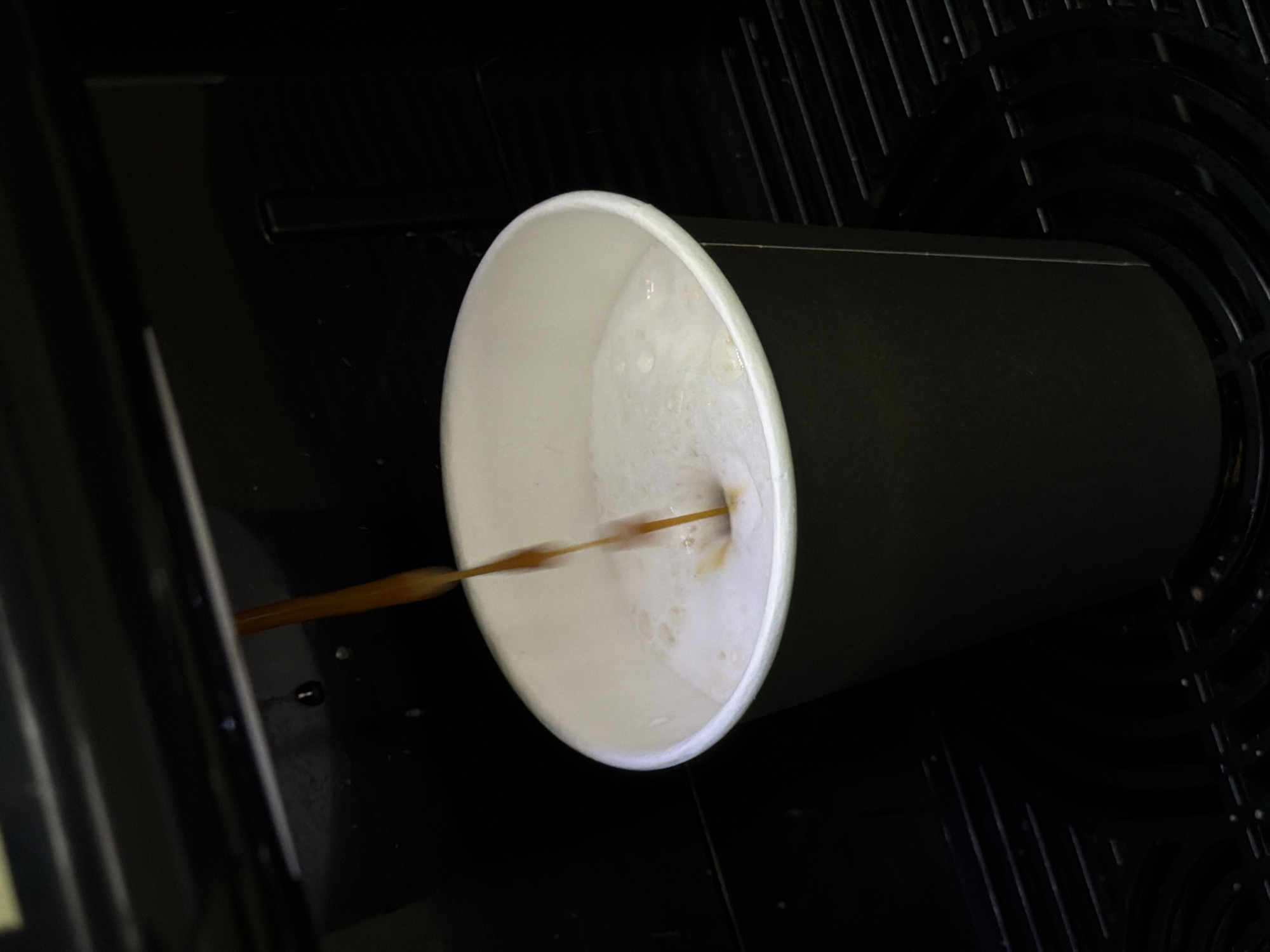 The image presents a close-up, high-angle view of a black paper coffee cup positioned within an automatic beverage dispenser. A thin, dark brown stream of liquid, likely coffee or espresso, is actively pouring into the cup from the left, indicated by slight motion blur. The cup is already partially filled with a light, creamy, and foamy beverage, suggesting a milky coffee drink such as a latte or cappuccino.
The immediate surroundings of the cup are dark and appear to be the interior mechanism of a coffee machine, featuring dark, possibly plastic or metal grates and ridges that form part of the machine's drip tray or internal structure. The lighting focuses on the inside of the cup and the pouring liquid, creating a stark contrast with the dark background. No people are visible, and there is no discernible text within the frame. The scene captures the moment of beverage preparation in an indoor setting, possibly in Chaikovsky, Russia. The time of day is not specified.
