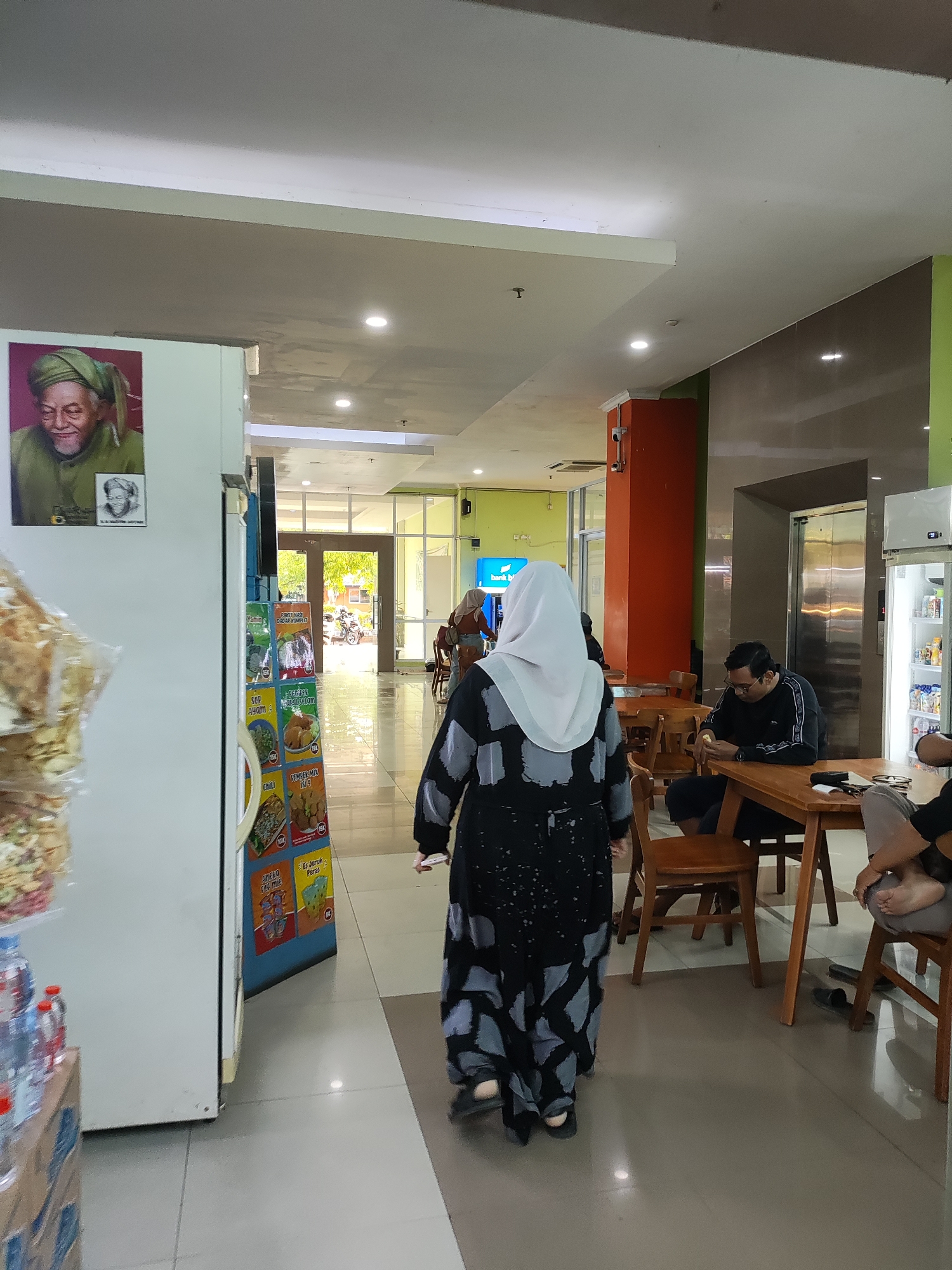 The image shows an interior scene, likely a public space with a cafe or dining area. A woman wearing a black and gray patterned dress and a white hijab is walking away from the camera. To her left, a refrigerator is visible, with a portrait of an elderly man in a green turban displayed on its side, alongside smaller framed images. The refrigerator is next to a display of various food products, possibly snacks or instant meals.
In the background, several individuals are seated at wooden tables. One man in a black long-sleeved shirt and glasses is looking down at his phone. Another person, partially visible, is also seated at a table. A large elevator door is visible behind the seated individuals. The environment appears to be indoors with bright lighting. The style of the portrait on the refrigerator and the presence of the elevator suggest it might be within a building like a hospital, school, or a public administrative complex. Given the cultural context suggested by the hijab and the portrait, the scene could be in Indonesia. However, without further information, it is not possible to pinpoint the exact location within Cirebon, Indonesia.