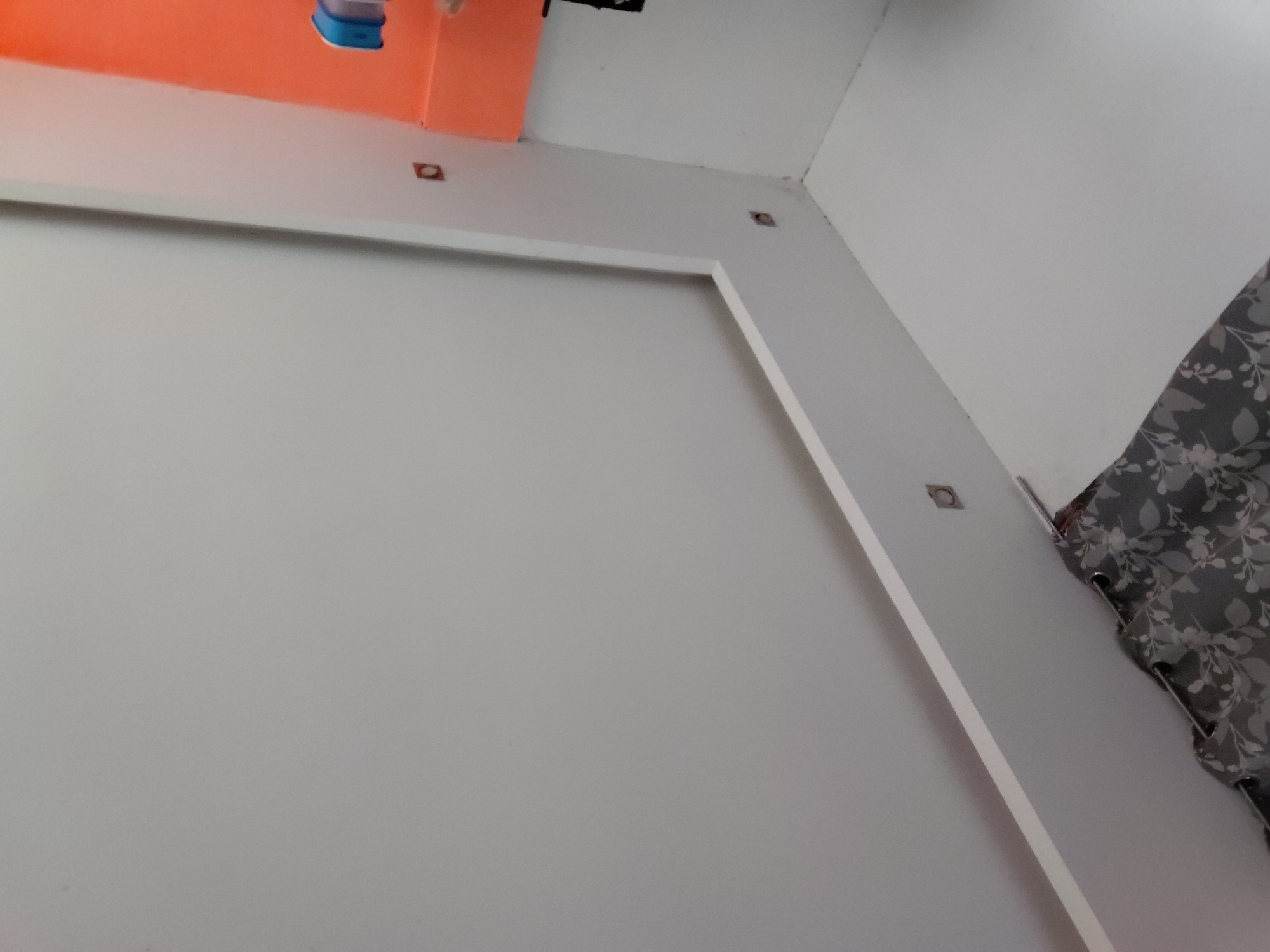 This image presents an interior view of a room, focusing on a corner where two walls and the ceiling meet, likely situated in Santa Ana, Venezuela. The walls and ceiling are painted in a light, possibly white or off-white, color, creating a clean and bright aesthetic. A distinctive white trim or molding outlines a rectangular section on the main wall, suggesting a decorative panel or a built-in feature.
In the upper left of the frame, a segment of a bright orange surface is visible, possibly part of an adjacent wall, a soffit, or a large fixture near the ceiling. A small, partially visible blue object rests on or is attached to this orange area. Scattered along the top edge of the white walls, close to the ceiling line and particularly at the corner, are several small, rectangular, dark brown or reddish-brown fixtures, which appear to be mounting brackets or anchors.
On the right side of the image, a patterned curtain is partially seen. It features a dark gray fabric adorned with a lighter-colored floral or leaf motif. The top edge of the curtain clearly shows metallic grommets, indicating it is hung from a curtain rod. The lighting within the room appears uniform, suggesting either diffused natural light or interior artificial lighting. There are no people, explicit activities, or visible text in the scene.