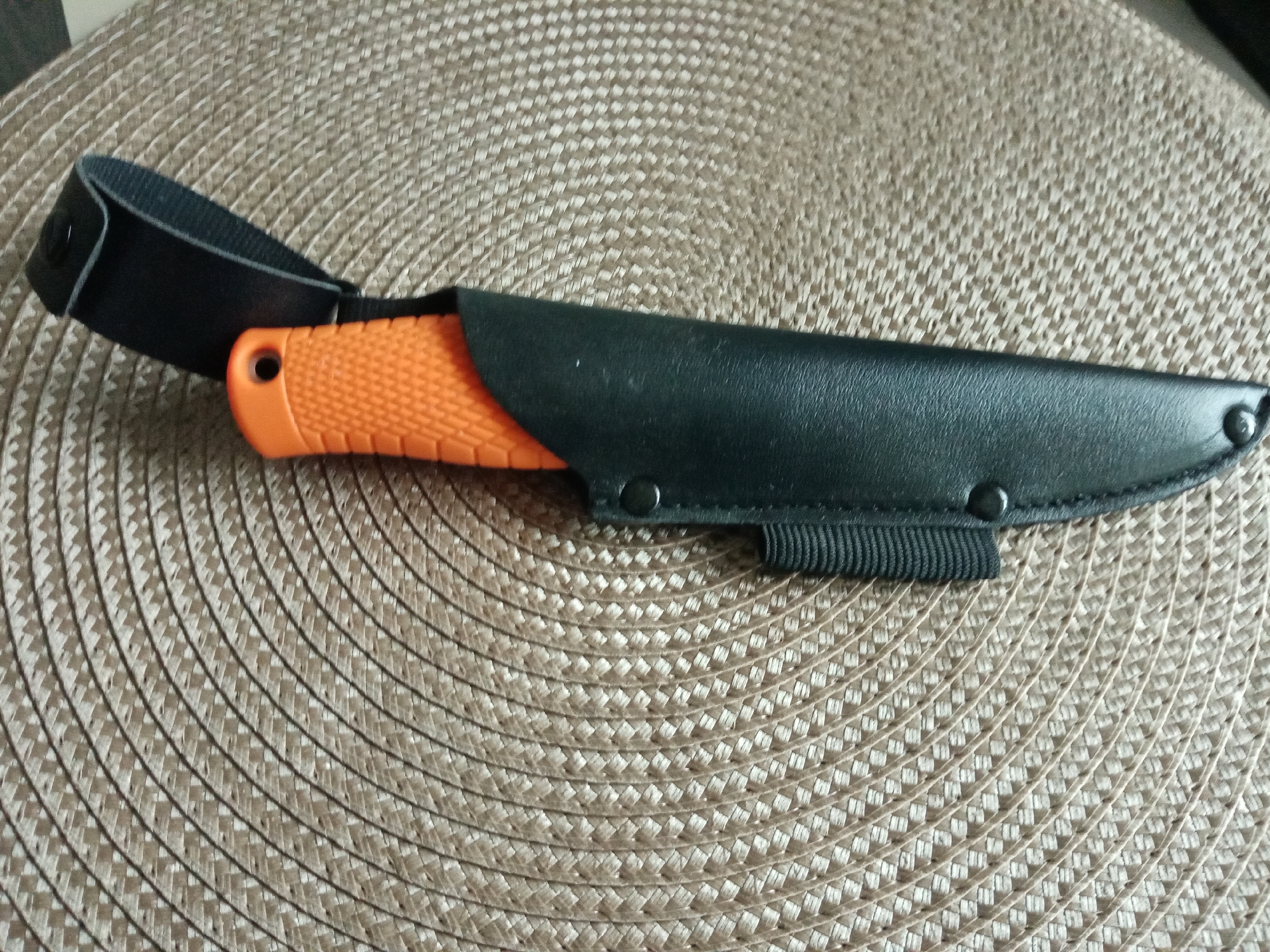 An orange-handled knife is sheathed in a black leather sheath on a tan, woven surface. The knife handle is visible, as is a black leather strap attached to the sheath. The sheath has black snaps. No other details suggest this image was taken in Elektrostal, Russia.