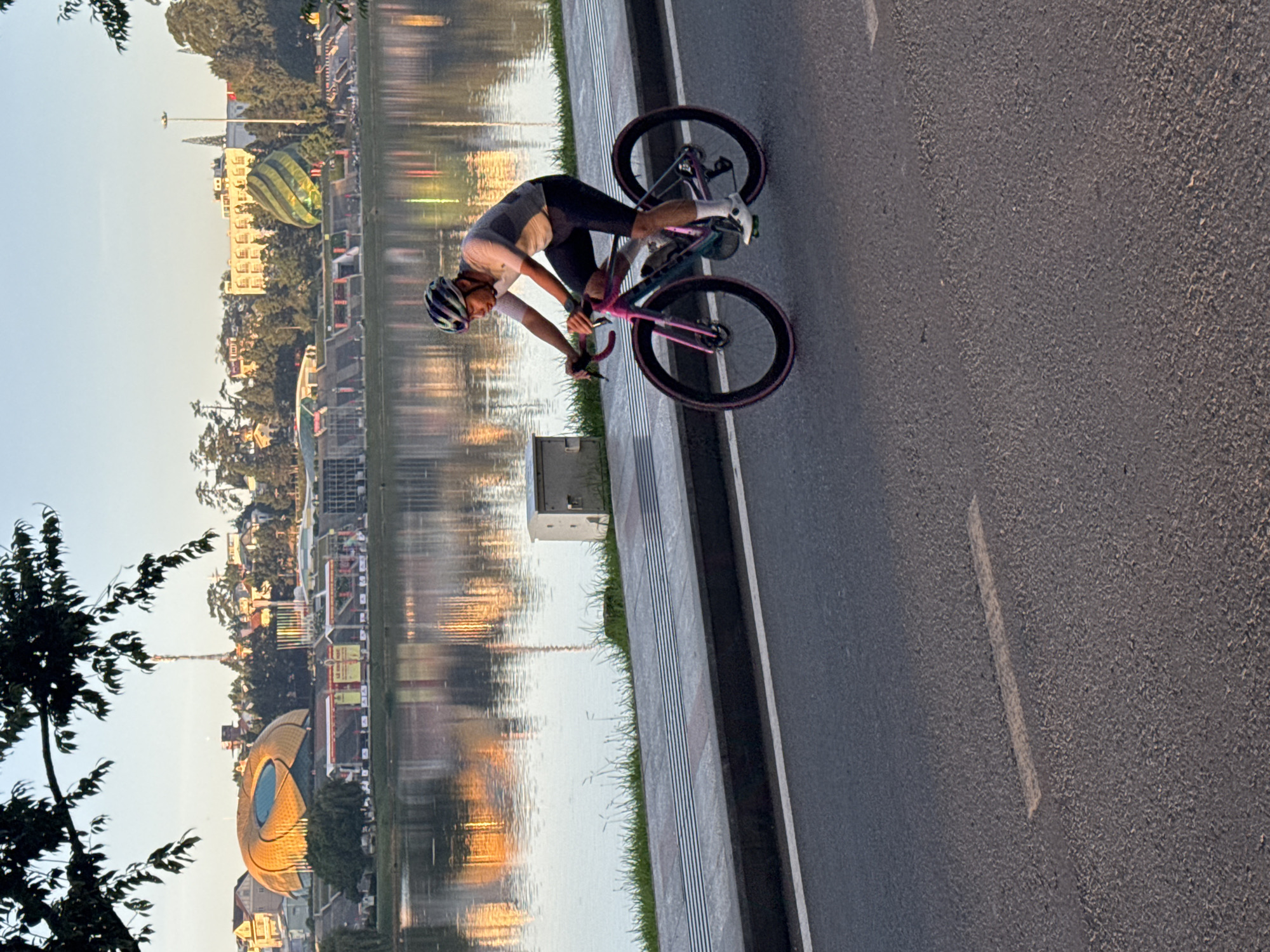 A lone cyclist wearing a helmet, a light-colored short-sleeved jersey, and dark shorts rides a pink and black bicycle on a paved road. The cyclist is positioned in the right-middle of the frame, with the bicycle in motion. The road features a white dashed line marking lanes. To the left of the road is a paved sidewalk with a textured surface and a curb. Beyond the sidewalk is a body of water, reflecting buildings and a clear sky. In the background, across the water, a city skyline is visible, featuring distinct architectural structures including a large, golden, domed building with a circular opening at its top, and a green, ribbed, balloon-shaped structure. Trees and other buildings are also part of the cityscape. The environment suggests a park or urban lakeside setting, likely in the vicinity of Xuân Hương Lake in Đà Lạt, Vietnam, given the architectural landmarks. The lighting indicates it is either early morning or late afternoon, with warm tones from the sun reflecting on the water and structures.