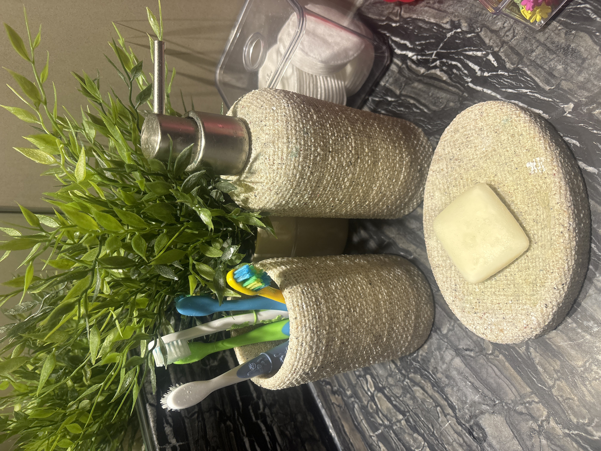 The image depicts a close-up, eye-level shot of a bathroom countertop. On the left, a textured cylindrical holder contains four toothbrushes of varying colors: white, green, blue, and yellow with colorful bristles. To the right of the toothbrushes, a tall, textured dispenser with a silver pump mechanism stands prominently. Behind these items, a lush green artificial plant with thin leaves is partially visible. In the foreground, a round, textured soap dish holds a light-colored bar of soap. To the right of the dispenser, a clear plastic container is filled with white cotton pads. The countertop itself has a dark, marbled pattern. In the lower right corner, a small clear container holds colorful small items that appear to be decorative pills or beads. The lighting suggests an indoor environment, likely a bathroom, with a reflective surface behind the plant, possibly a mirror. There are no people or animals present in the scene. The image does not provide any specific indicators of its location in Dubai, United Arab Emirates.