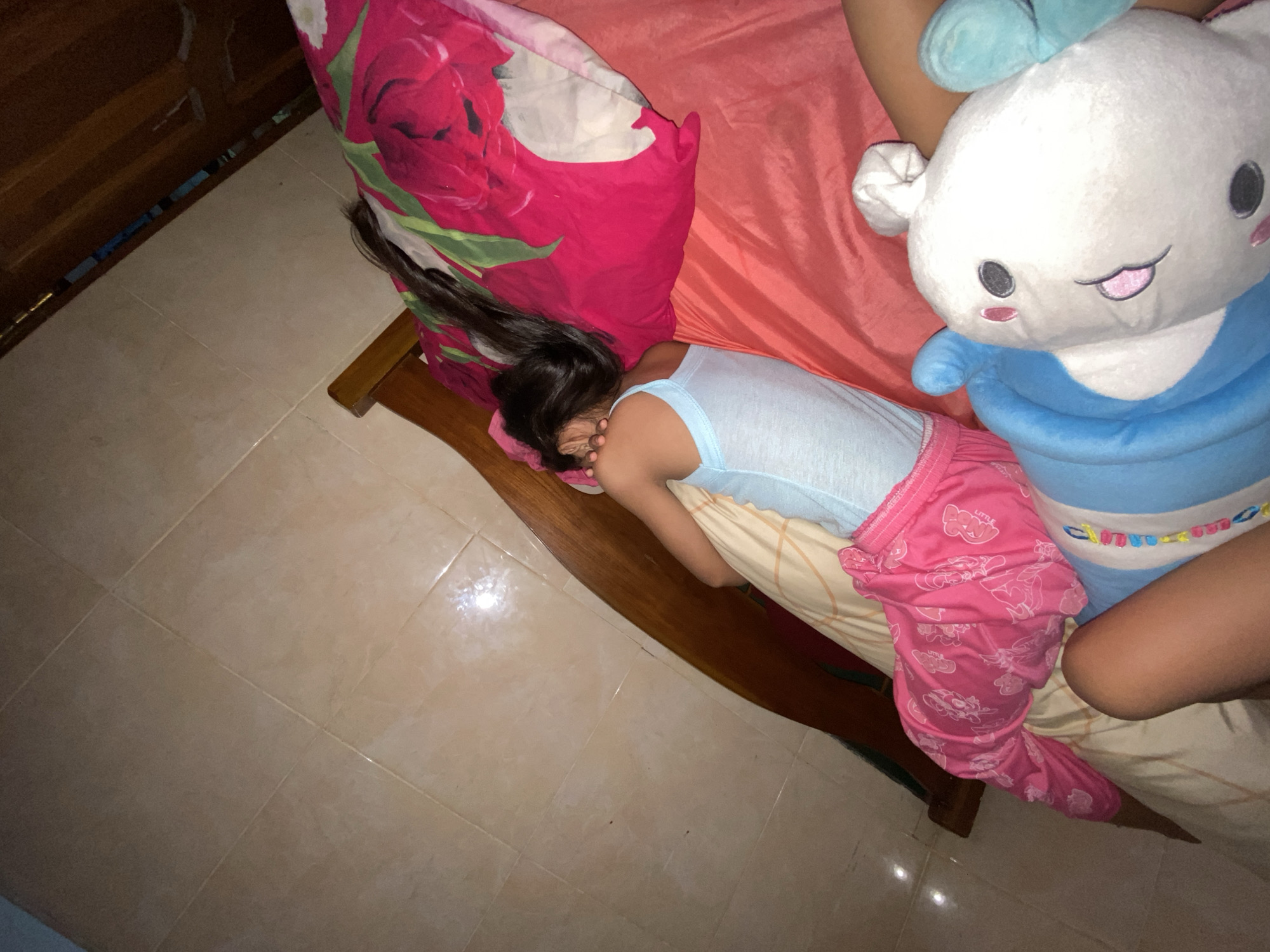 A child is sleeping on a bed with a wooden headboard in Gelam, Indonesia. The child is wearing a light blue tank top and pink pajamas with a cartoon pattern. A large stuffed animal, primarily blue and white with some pink accents, is lying on the bed next to the child. The bed is covered with a pink floral patterned sheet and a red sheet. The floor next to the bed is tiled in a light tan color.