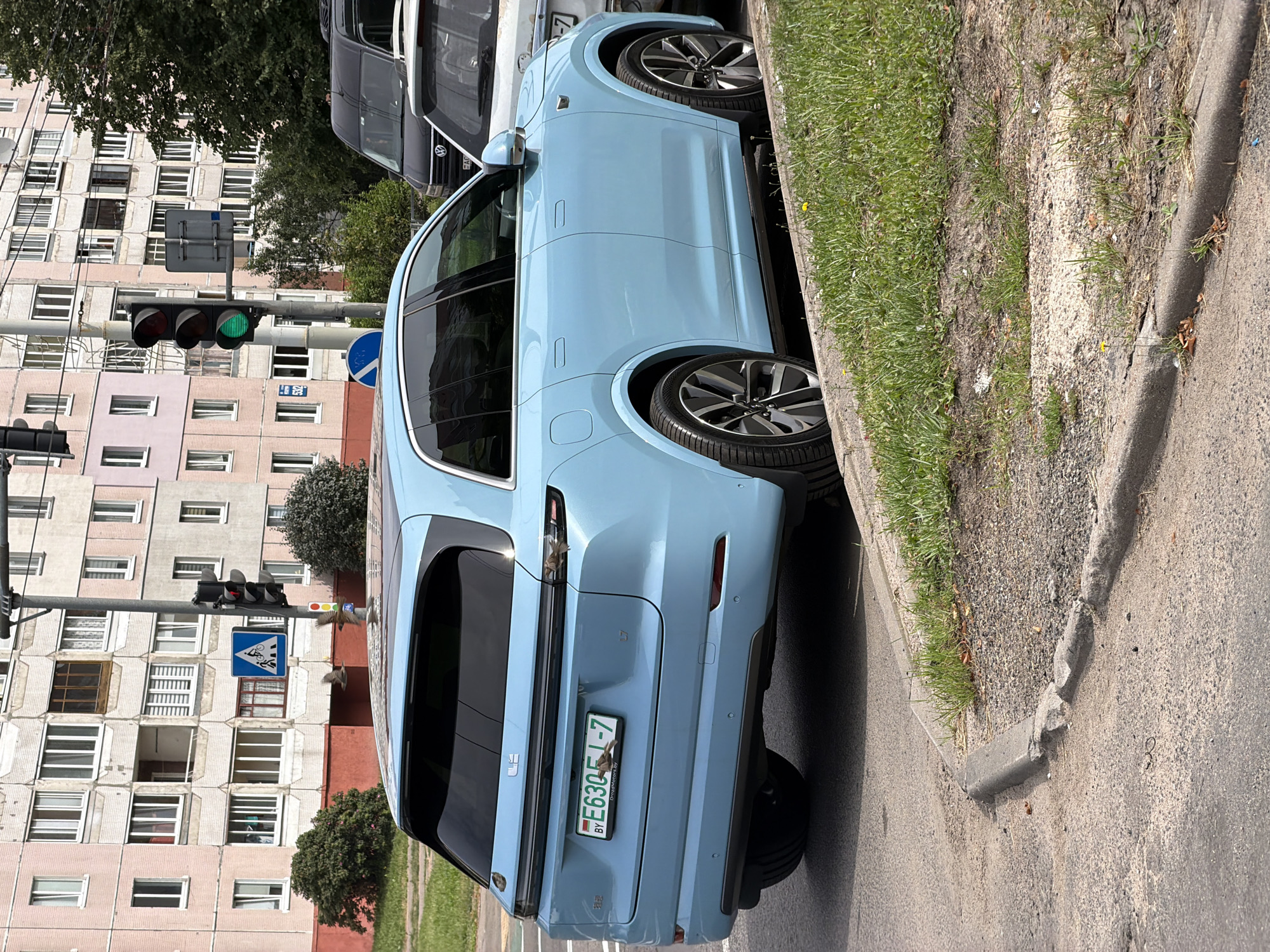 A pale blue electric SUV, identified as an L7 and bearing a Belarusian license plate E63051-7, is parked at the side of a street in Minsk, Belarus. The vehicle is positioned next to a grassy embankment with a concrete curb. In the background, a multi-story residential building with pink and beige facades and numerous windows is visible. A traffic light, currently showing a green signal, is also present, along with a blue pedestrian crossing sign depicting a person on a bicycle. Several small birds are perched on the rear of the car. The street appears to be an urban setting, likely in a residential area of Minsk.