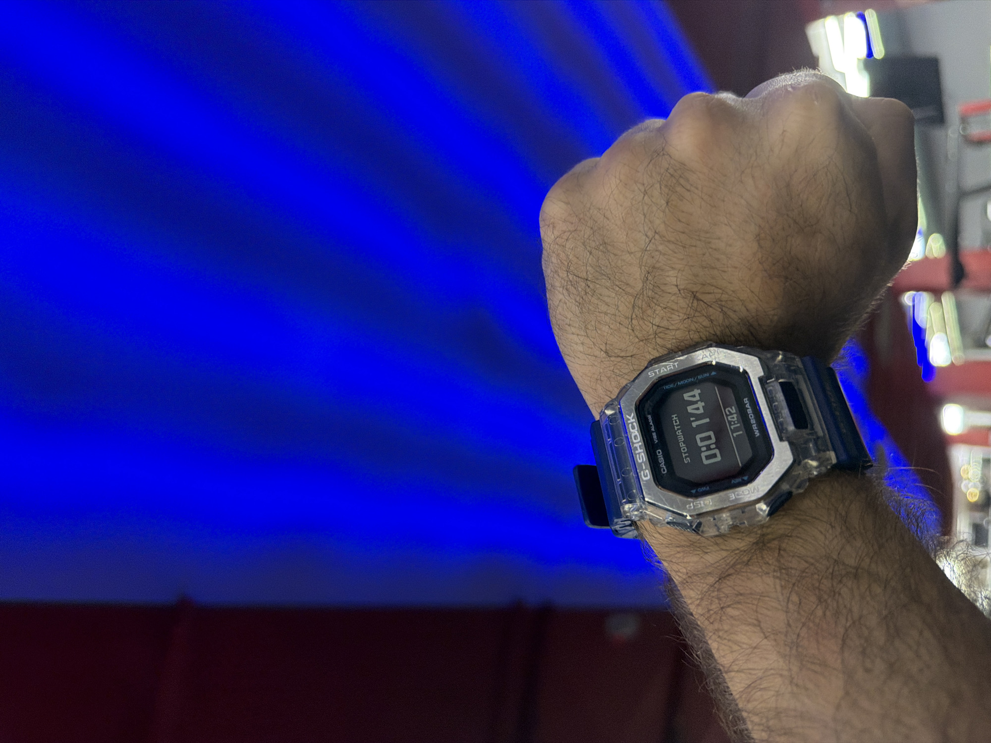 A wrist with a G-Shock watch is prominently displayed in the foreground, against a backdrop featuring a striking blue and red lighting effect. The watch displays a timer reading "0:0 1'44" and the time "11:42." The background also contains blurred elements suggesting a somewhat cluttered environment with some indistinct red and white shapes. The location in the city of Krasnoyarsk, Russia, is not discernible from the image.