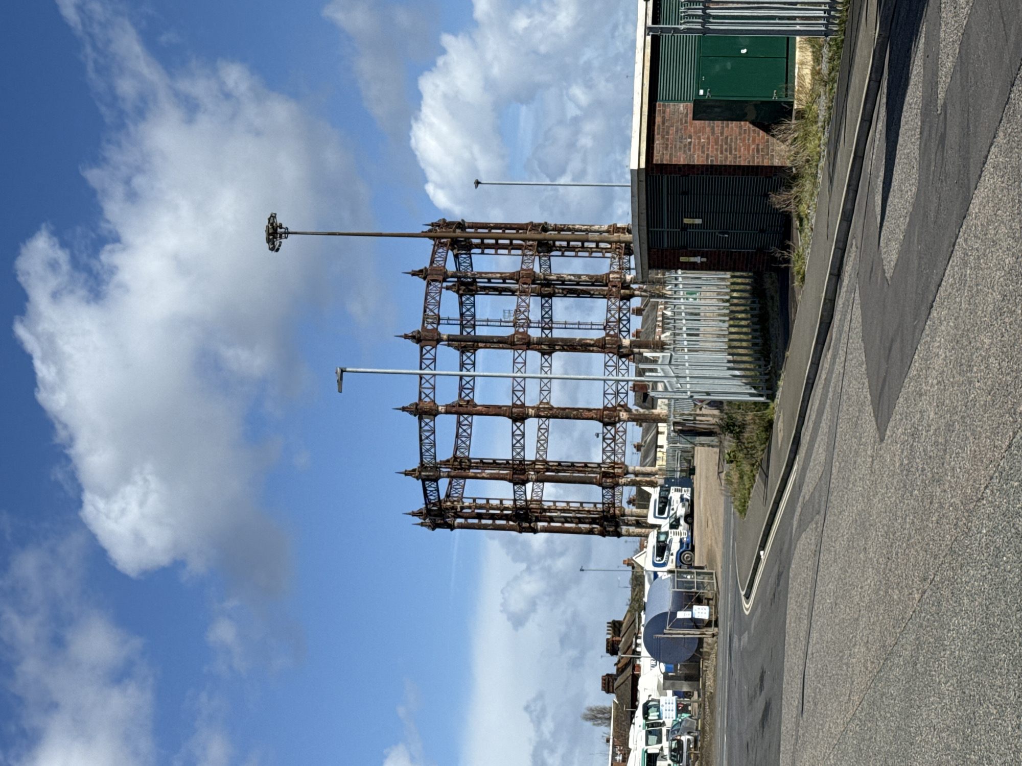 The image depicts an outdoor, urban-industrial scene in Gorleston-on-Sea, United Kingdom, under a blue sky with scattered cumulus clouds.
The central and dominant subject is a large, derelict gasometer frame, heavily rusted, with a multi-tiered, cylindrical lattice structure composed of vertical uprights and diagonal cross-bracing. The top of the frame features ornate, pointed finials.
In the mid-ground to the left of the gasometer, an industrial yard is visible, containing several white and blue commercial tractor units, a silver passenger car, and a large, blue, cylindrical storage tank. This area is partially enclosed by fencing. Behind the industrial yard and the gasometer, rows of traditional terraced or semi-detached residential houses with red-tiled roofs are present.
The foreground features an asphalt road with a visible curb and double white lines, curving from the lower left towards the right. On the right side of the road, a silver metal palisade fence encloses a small, single-story utility building. This building has a brick base and dark green louvred panels for its upper section and doors. Patches of grass and weeds are visible along the roadside and around the utility structure.
Three distinct streetlights are present: a standard streetlamp to the left of the gasometer, a taller high-mast lighting pole with a circular cluster of fixtures positioned behind the gasometer, and a slender streetlamp further to the right.
No individuals are visible. The scene is static, illuminated by natural daylight, casting moderate shadows.