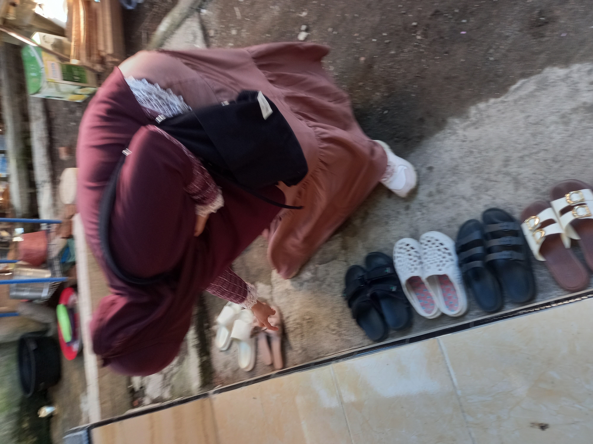 A woman wearing a maroon hijab and a long, dusty rose dress is bending down to pick up a pair of pink slides. She is outdoors, standing on a concrete floor in what appears to be a residential area. In front of her are several pairs of shoes, including black slides and white slides with a patterned footbed. To her left, the floor transitions to lighter colored tiles, with a black bucket and a red plate with a green object on it visible in the background. The scene is likely in Tempos, Indonesia, given the context provided.