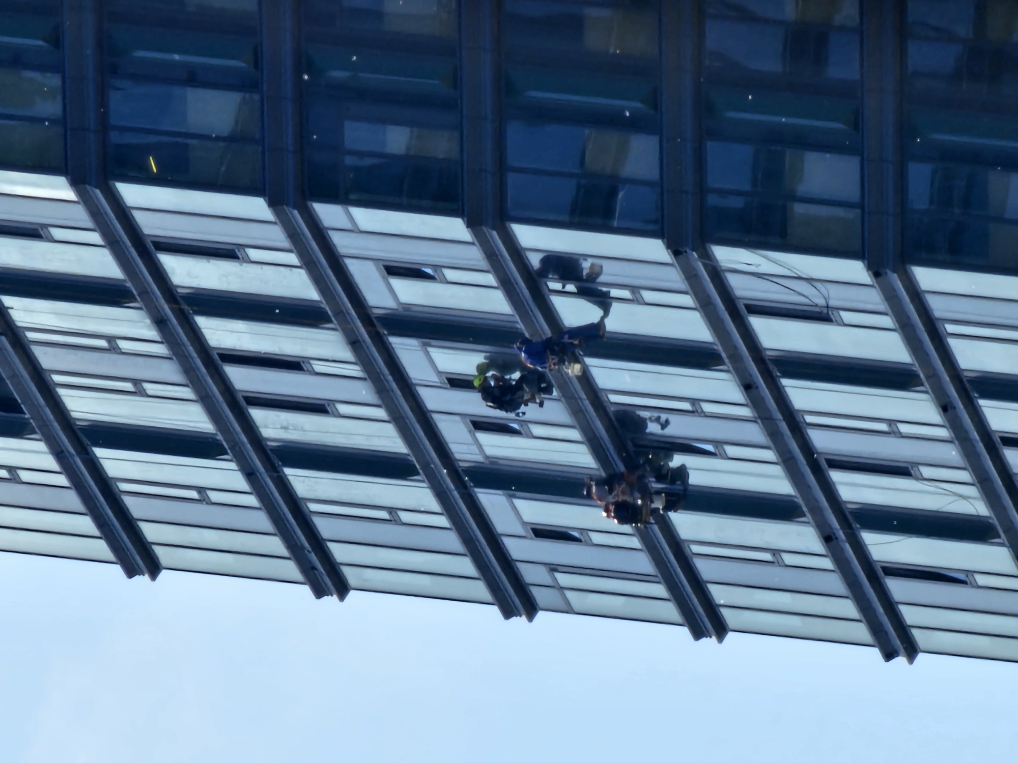 The image displays a modern high-rise building facade in Moscow, Russia, featuring a striking pattern of dark blue glass panels and light grey/white horizontal sections. The perspective is unusual, suggesting a view from below looking up, or a rotated image, with a clear blue sky occupying the lower part of the frame.
The central elements are the reflections visible on the dark glass surfaces. These reflections distinctly show at least two individuals, along with their equipment. One figure, visible on the left within the cluster of reflections, appears to be wearing a blue uniform and potentially a helmet, equipped with a harness or part of a suspended platform. A second figure is reflected to the right and slightly below the first, also equipped for work on the building's exterior.
The activity depicted through these reflections is exterior building maintenance, most likely window cleaning, with workers suspended from the building. The bright, clear blue sky indicates it is daytime with favorable weather conditions. The building's architecture is contemporary, characteristic of a large urban environment. There is no visible text in the image.