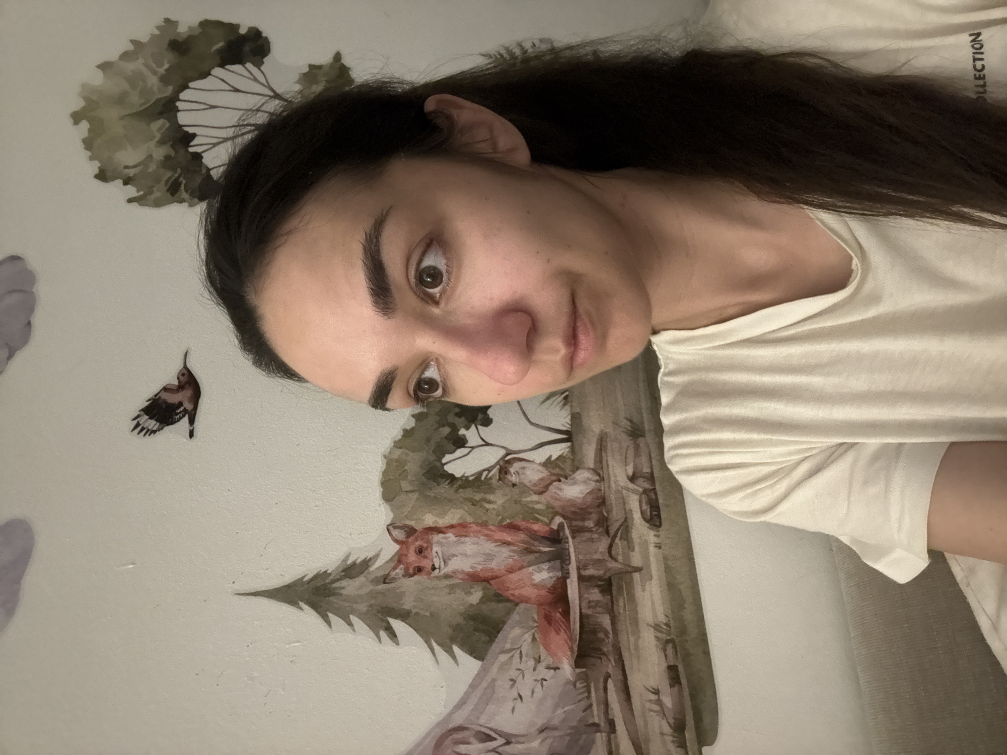 A woman with long dark hair is posing in front of a wall decorated with nature-themed decals, including trees, a fox, a bird, and other forest creatures. The woman is wearing a light-colored t-shirt. The photograph appears to be taken indoors, likely in a residential setting. There is no indication of a location within Bat Yam, Israel, or any other specific city.
