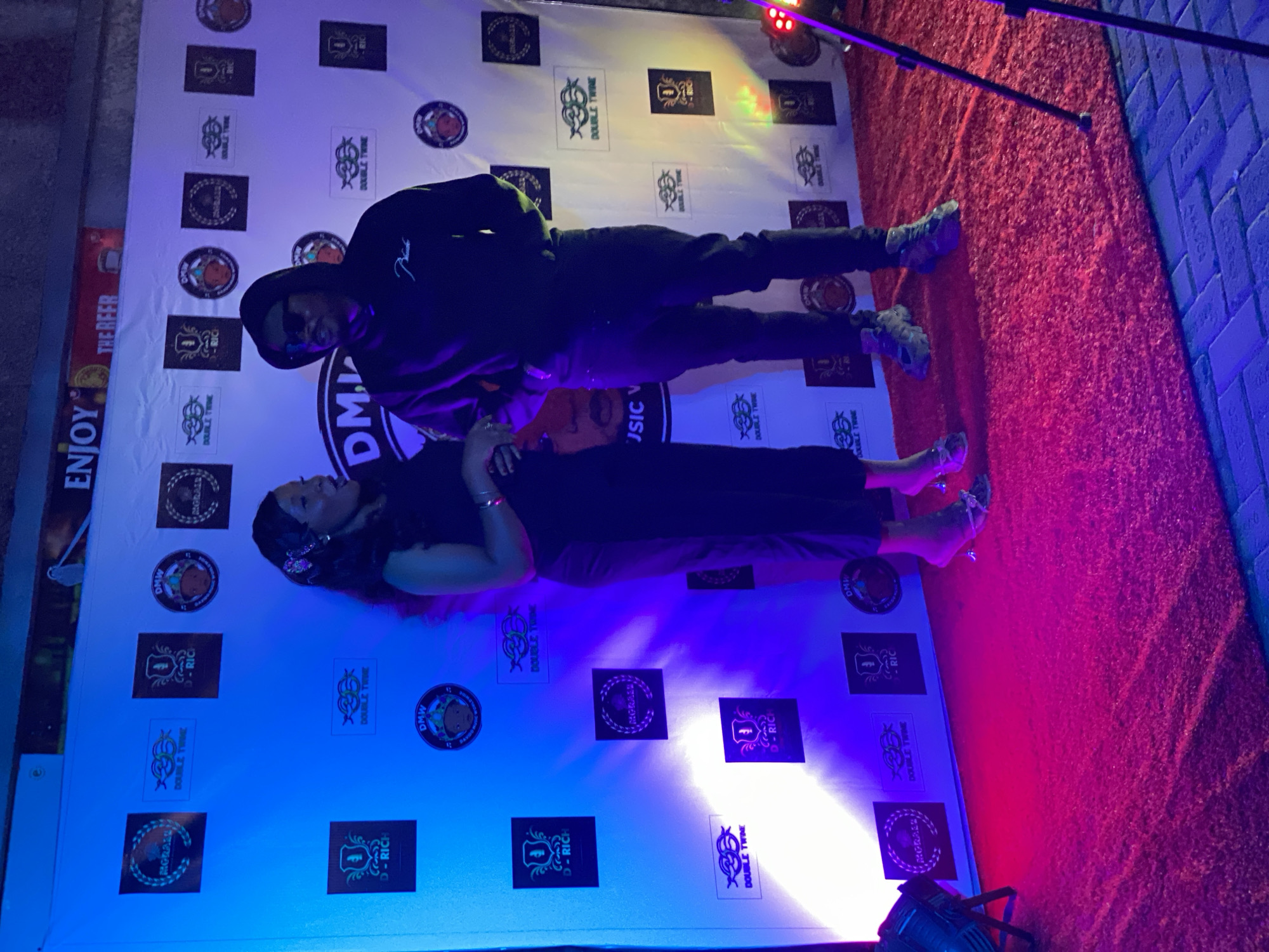 The image shows two individuals posing in front of a backdrop covered with logos, possibly at an event. A man in a dark hoodie and sunglasses is standing behind a woman who is lying down. The backdrop features various logo designs, including "DMW," "DOUBLE TWINE," and "D-RICH." The scene appears to be illuminated with blue and red lighting. A red carpet extends beyond the backdrop, and some equipment, such as light stands, is visible. The OCR mentions "Iranla, Nigeria," suggesting this is where the event took place.