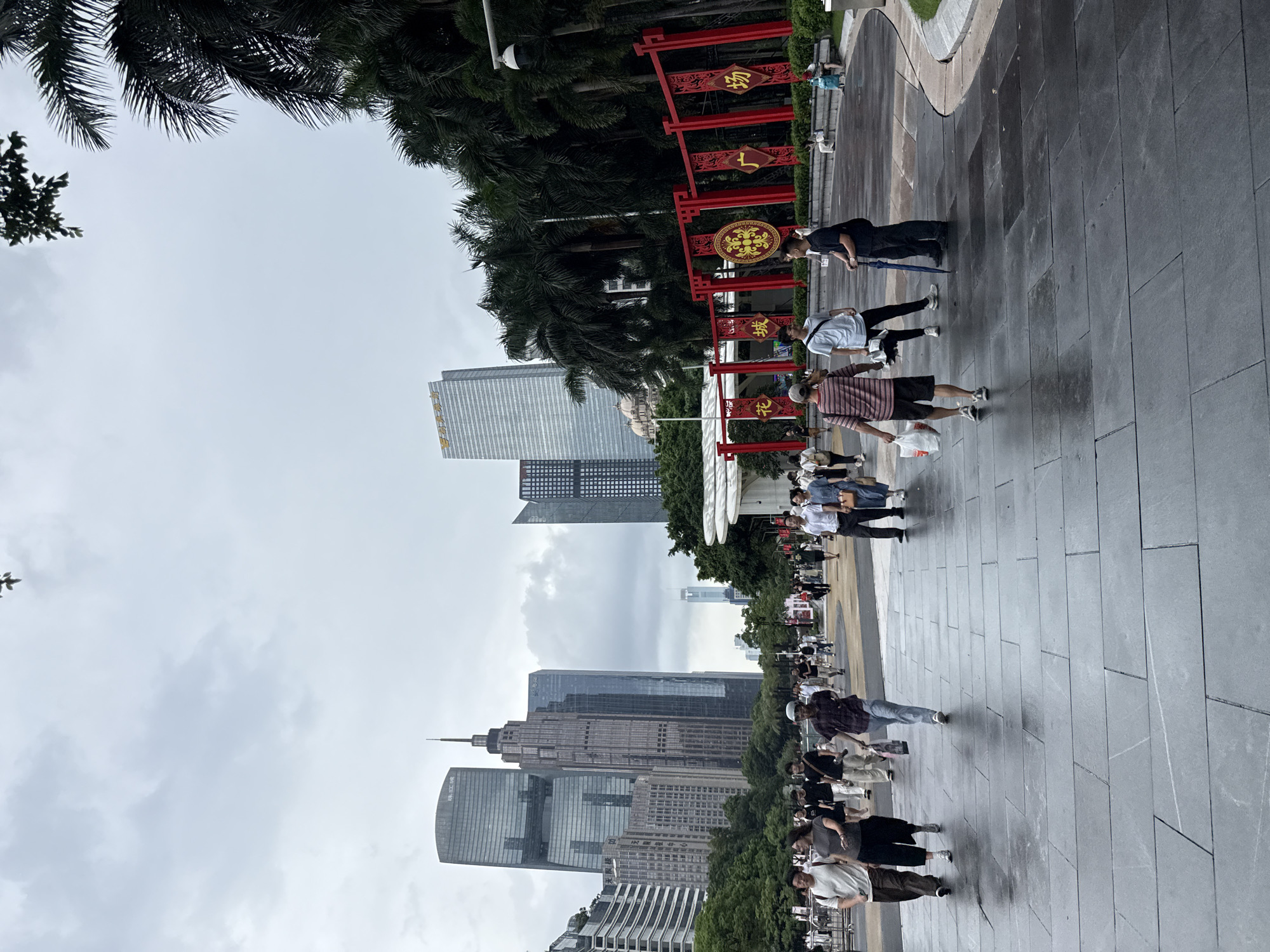 The image depicts a wide, wet pedestrian walkway in Tianhe District, China, likely after a rain. Tall, modern skyscrapers form the backdrop to a streetscape dotted with people. In the foreground, several individuals are walking, some carrying shopping bags. The pavement is made of large, dark gray rectangular tiles that reflect the overcast sky. To the right, lush palm trees are visible, framing a red decorative archway with Chinese characters. A security camera is mounted on a pole near the palm trees. Further back, a group of people are seated on a low wall or bench. The overall atmosphere is that of a bustling urban public space on a cloudy day.