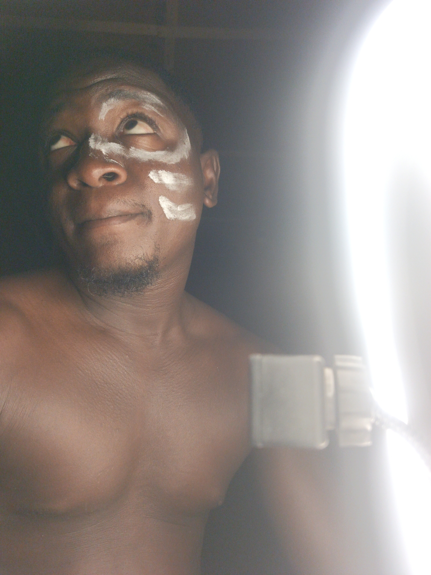 A close-up shot reveals a shirtless Black man with white face paint near his eyes and on his nose. He is looking upwards and to the left. The background behind him is dark, while a bright light is on his right, partially obscuring the view. Part of what appears to be a device is visible between the man and the light source. The location and city are unknown.
