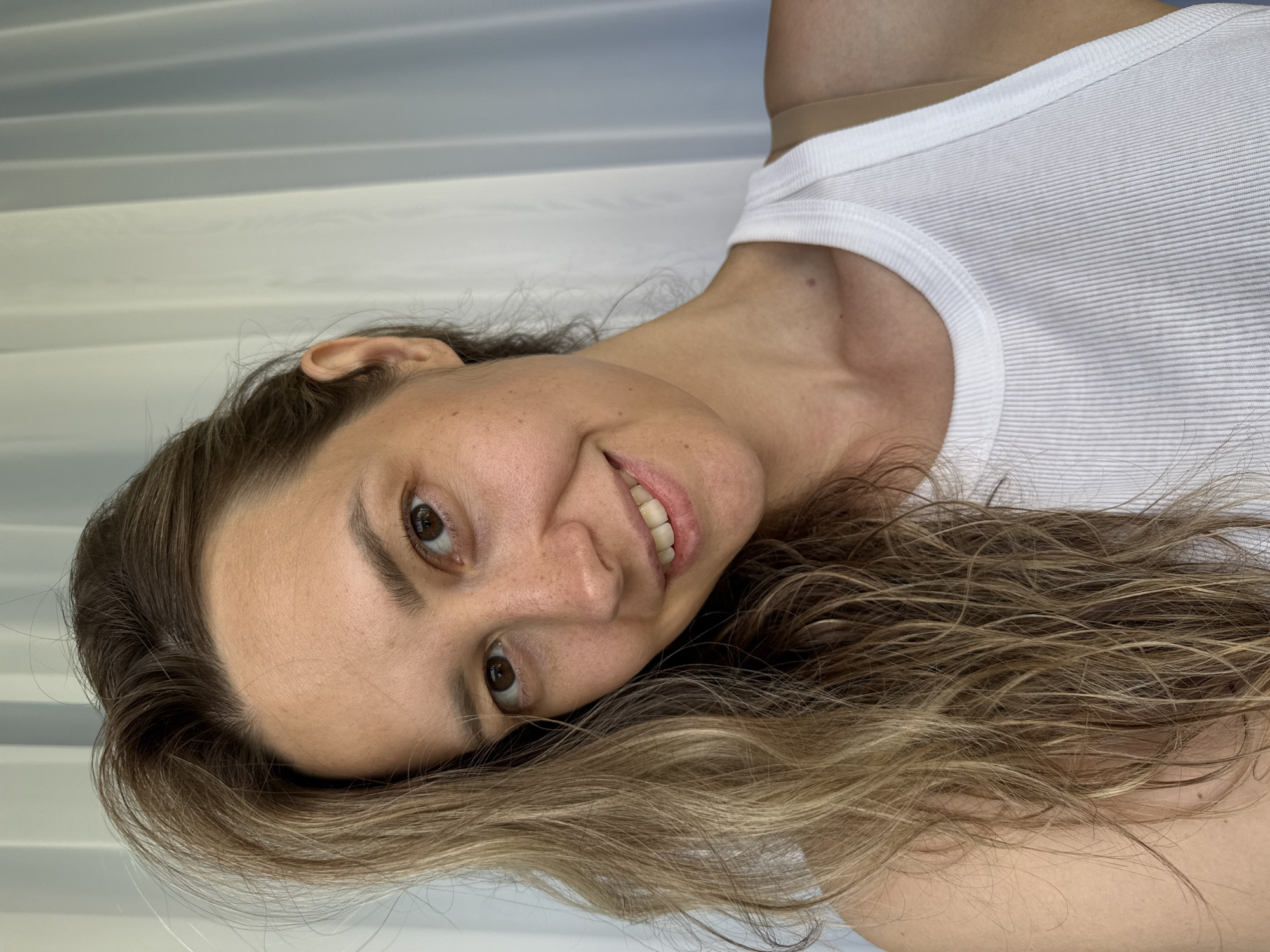 A medium-shot, selfie-style photograph shows a woman with shoulder-length, wavy blonde hair that is partially covering the left side of her face. She is wearing a white ribbed tank top. Her head is tilted slightly to her left. She has light brown eyes, a light complexion with freckles, and is smiling, showing her teeth. She is indoors, with a blurred white curtain in the background, suggesting a location like a hotel room or an apartment in the city of Nebug, Russia. The overall lighting is bright and natural.