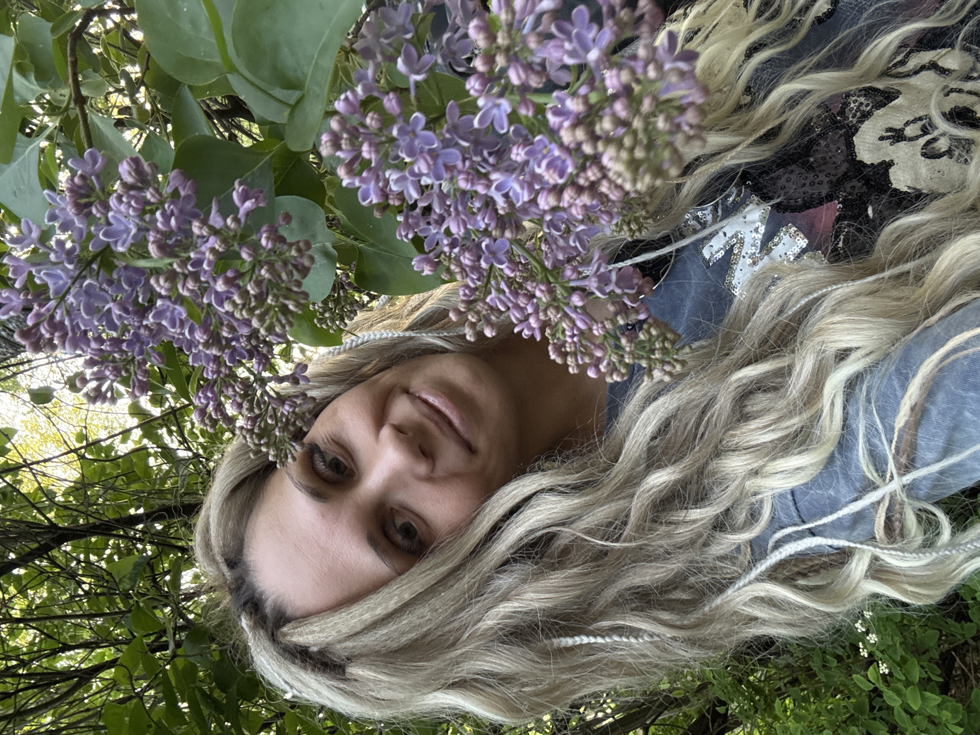 A woman with long, wavy blonde hair poses with a lilac bush. The woman is wearing a blue garment and a top with sequins. She is lying on her side, looking upwards, possibly on the ground in an outdoor setting. The lilac bush is in full bloom, with clusters of purple flowers. The background includes green foliage and dappled sunlight. The location is specified as Rusanovo, Russia.