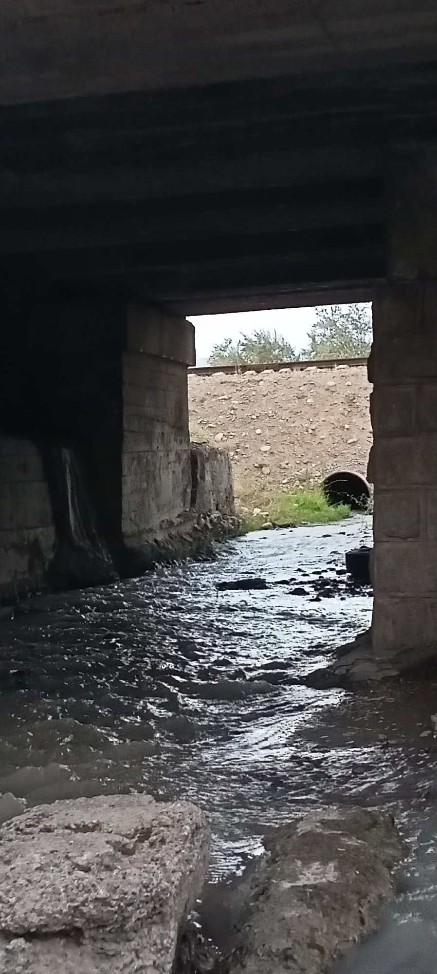 The image shows a view from under a concrete bridge, looking out towards a wide, shallow stream flowing through a dry, rocky landscape. The water is murky and appears to be carrying debris, with ripples and waves indicating flow. On the left side of the frame, a concrete wall of the bridge descends to the water's edge. On the right, a similar concrete pillar supports the bridge. Beyond the stream, a sloping embankment of loose soil and rocks is visible, topped by a weathered stone wall. In the distance, trees are sparsely scattered against a cloudy sky, and what appears to be a railway track runs along the top of the embankment. A dark, circular culvert is visible in the middle distance, partially obscured by grass. The overall impression is one of a somewhat neglected, utilitarian infrastructure area. This scene is located in Sokuluk, Kyrgyzstan.