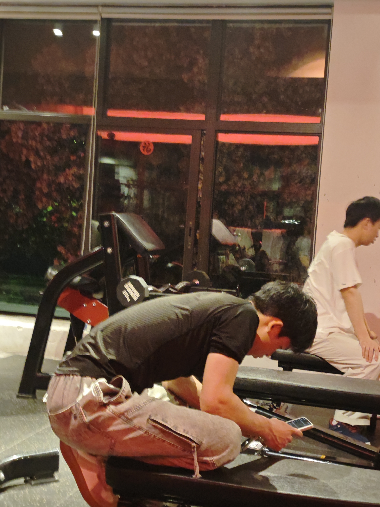 A photo shows a man in a black shirt and light-colored jeans kneeling on a black bench, holding a smartphone. The man is focused on the phone. Behind him, another man in a white shirt is seated on a bench. Gym equipment, including a weight rack and barbell, is visible. The setting appears to be a gym, as indicated by the equipment. Large windows display an outdoor scene at night. A red light strip is seen through the window. The location is Chongqing High-tech Zone, China.
