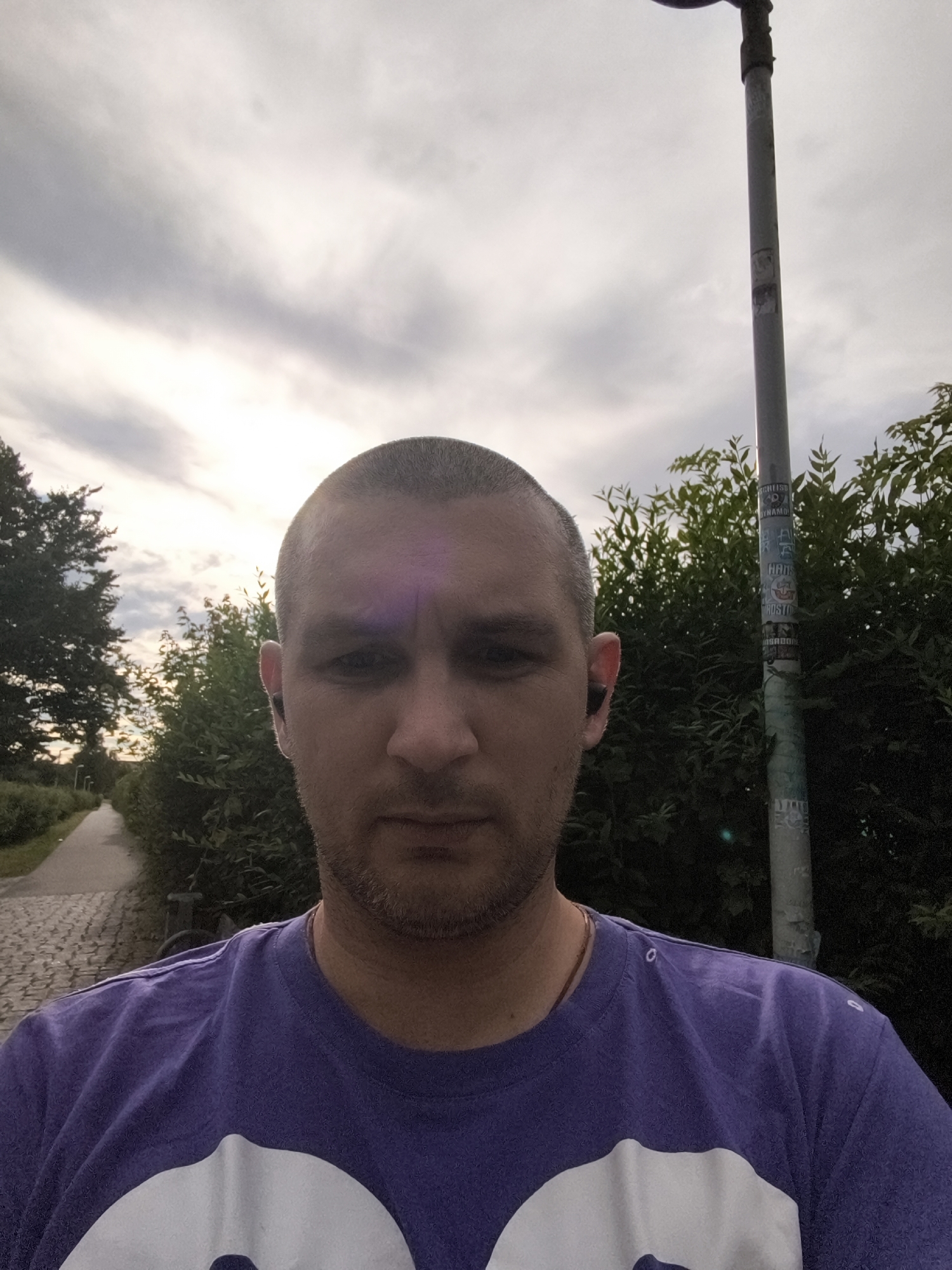 The image shows a man taking a selfie outdoors, with a paved path and trees in the background, characteristic of a park or recreational area, possibly located in Wolgast, Germany. The man is wearing a purple t-shirt and earbuds. The sky is cloudy with patches of sunlight. A lamppost with stickers is visible to the right of the man.