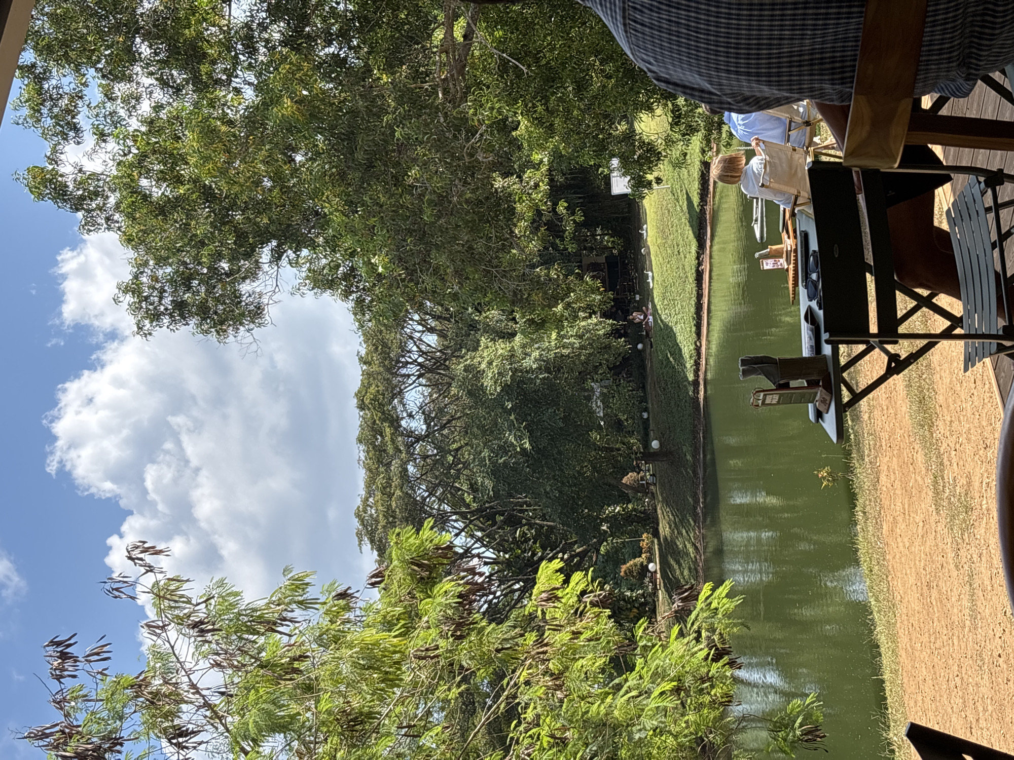 This outdoor, horizontal-oriented image depicts a serene scene at what appears to be a resort or dining establishment in Kuta Selatan, Indonesia. The dominant feature is a calm, green body of water, possibly a pond or canal, running horizontally across the middle ground. Lush green trees and dense foliage line the far bank and extend into the background, providing ample shade. The sky above is blue with scattered fluffy white clouds, indicating clear weather.
In the immediate foreground on the right, part of a wooden deck or platform is visible, supporting what looks like rustic wooden chairs and a table. Closer to the camera, a person's upper body wearing a blue plaid shirt is seen, suggesting they are seated at one of these tables, looking out towards the scene.
Further into the middle ground, alongside the water, two individuals are seated on black folding chairs. One person, with light-colored hair, faces away from the camera, looking towards the water. A small table or tray in front of them holds sunglasses and possibly a menu or information card.
The banks of the water are covered in green grass, contrasting with a sandy or dirt path on the right side of the water. Decorative light fixtures or small lanterns are spaced along the far grassy bank. Several white objects, possibly small statues or decorative accents, are also visible on the distant grass. The dense, tropical vegetation, bright daylight, and casual seating suggest a relaxed leisure activity, likely dining or enjoying the scenic view. No discernible text is visible in the image.