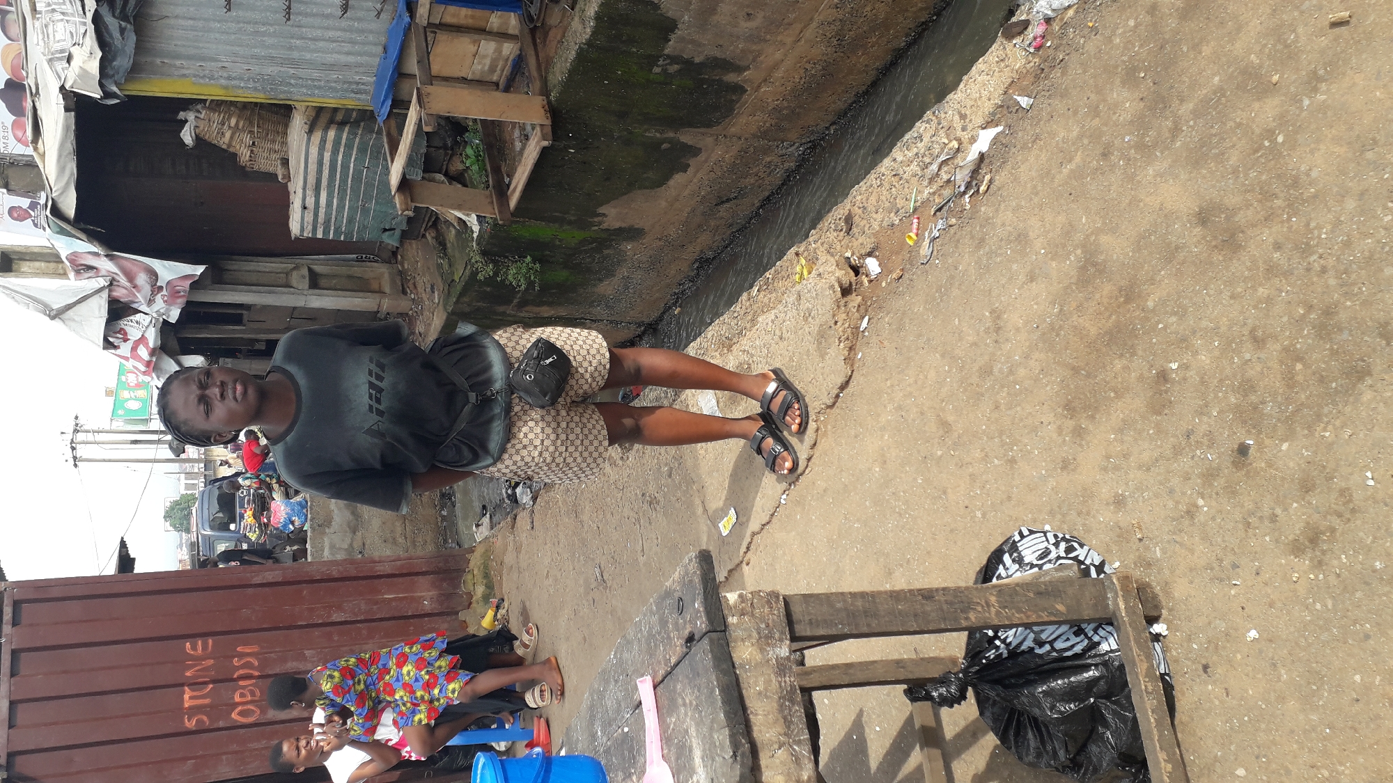 In Nkpor, Nigeria, a young woman stands on a dirt path next to a narrow drainage ditch. She is wearing a dark t-shirt with a graphic design and patterned shorts, with a small black pouch slung across her chest. Her feet are clad in black sandals. Behind her, a large, rust-colored container is marked with white lettering that appears to read "STONE OBOSI". Further in the background, a cluttered marketplace is visible, with various stalls and people engaged in commerce. The overall environment suggests a busy, outdoor market area, potentially in a commercial district of the city.