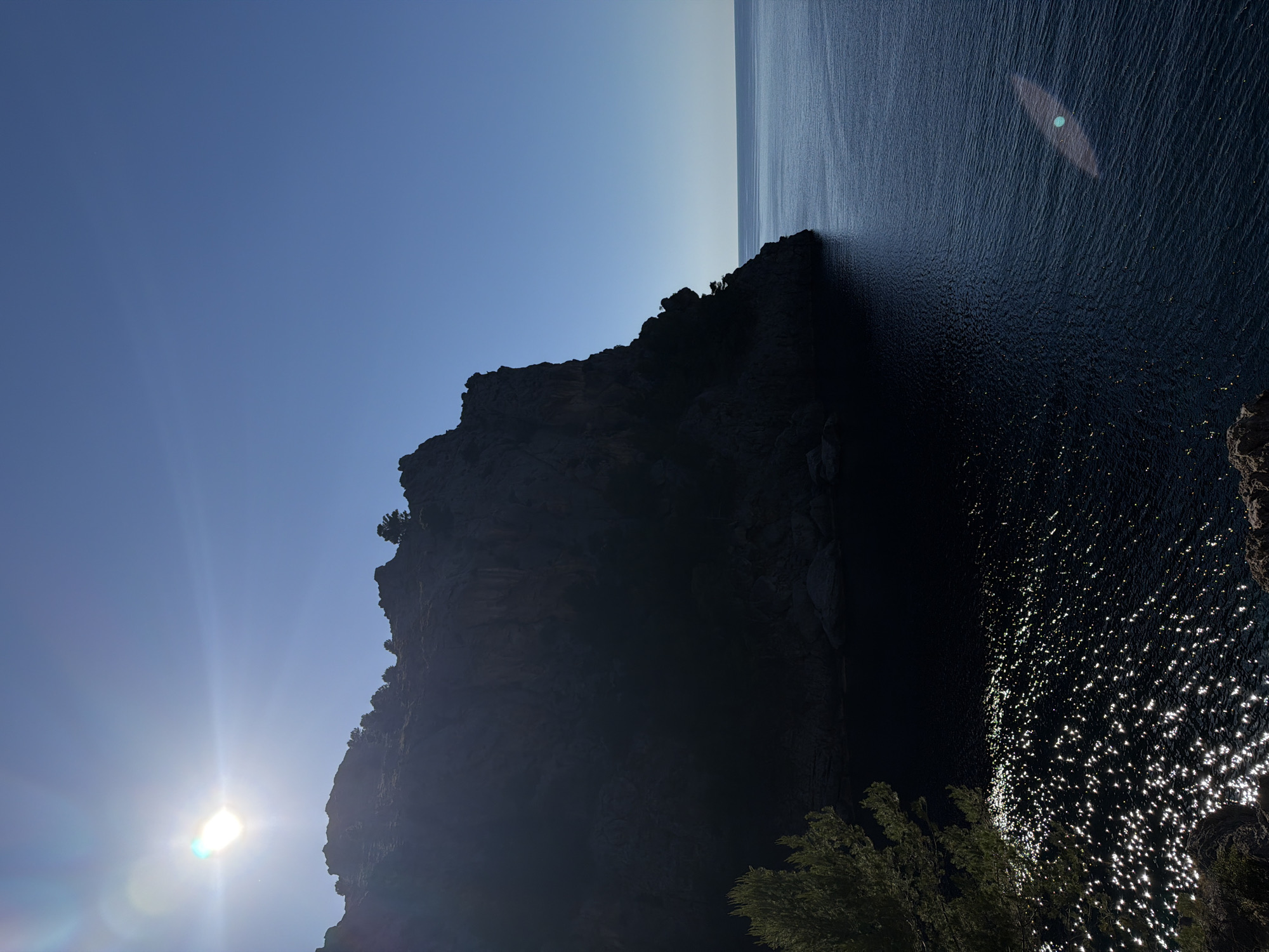 The image depicts a steep, rocky cliff face descending into the dark waters of the Mediterranean Sea, likely near Escorca, Spain, given the request. The sky transitions from a bright, pale blue at the horizon to a deeper blue higher up. The sun is visible, positioned low in the sky, creating a bright glare in the lower left corner of the image. Sunlight reflects off the surface of the water, creating sparkling patterns. A few trees are visible near the base of the cliff. The cliff dominates the image, casting a large shadow over a significant portion of the water's surface.