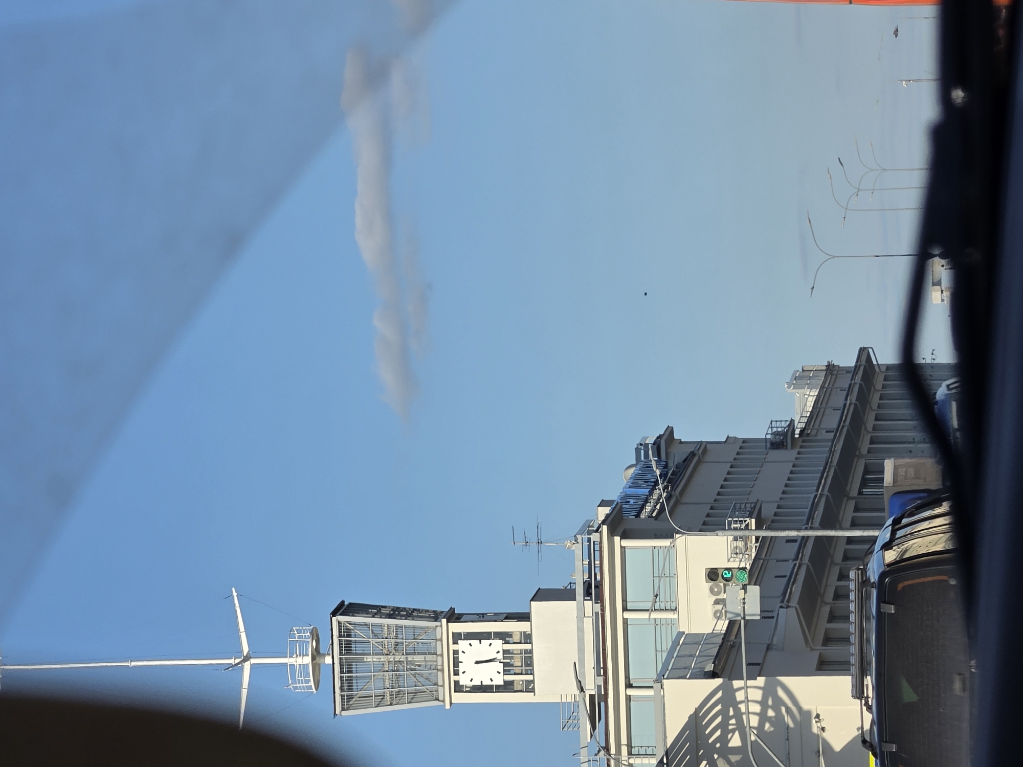 The image shows a street view in Nizhny Novgorod, Russia, focusing on a white building with a prominent clock tower. The clock on the tower shows the time as approximately 10:50. The building has multiple levels with windows and balconies. In the foreground, the roof and rear window of a dark-colored SUV are visible, suggesting the photo was taken from inside a vehicle. A traffic light displaying green is also visible to the left. The sky is blue with a few scattered clouds, and streetlights line the side of the road. The overall scene depicts an urban environment on a clear day.