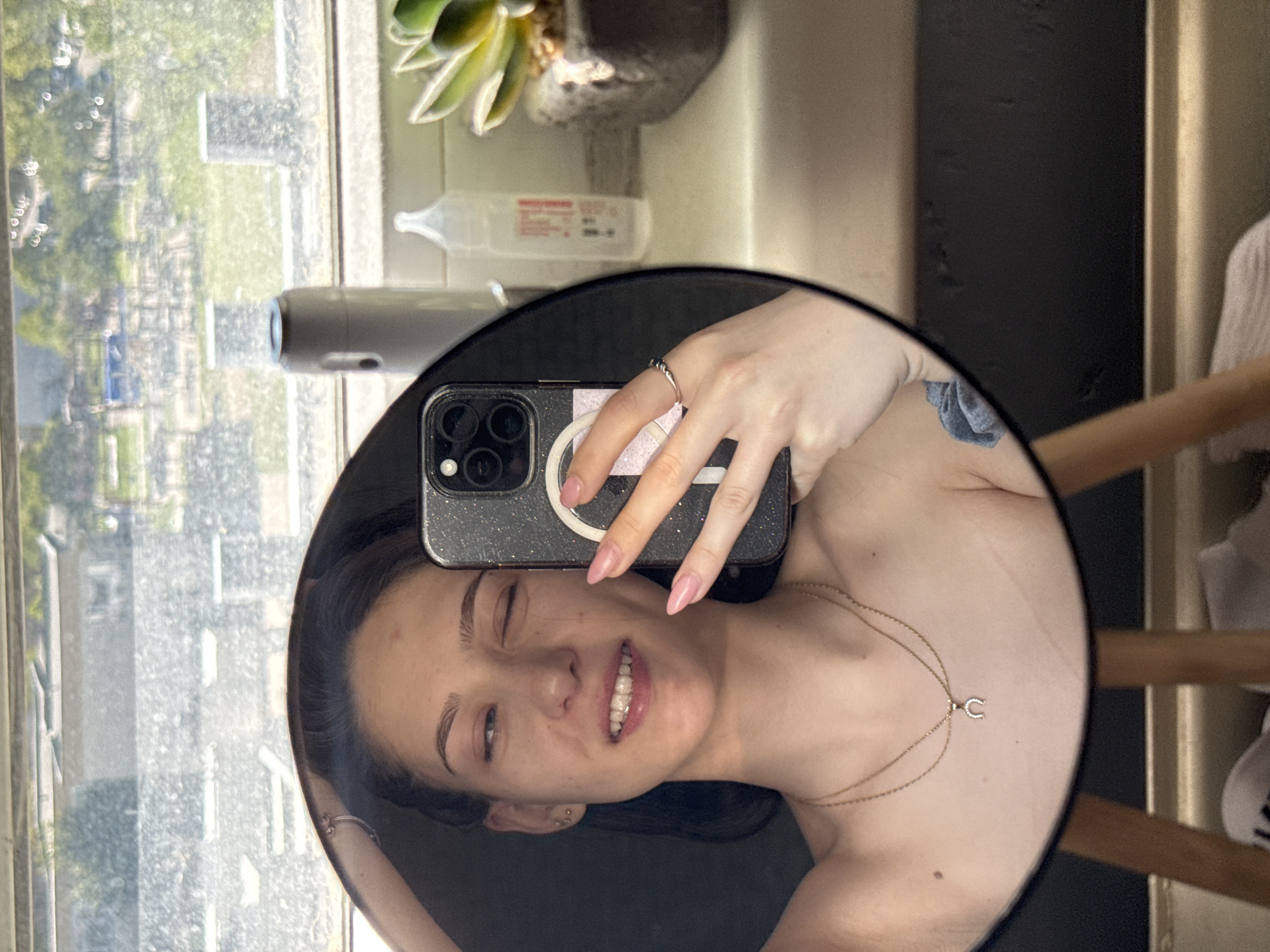 A woman is shown in a mirror selfie. She's holding an iPhone in her right hand, a ring visible on her ring finger. Her face is angled to the left. She wears gold jewelry and has a small tattoo on her right shoulder. The mirror is on a surface near a window, with a view of trees and buildings outside. Behind the mirror are objects including a plant and medical-type fluid. The location can not be determined.
