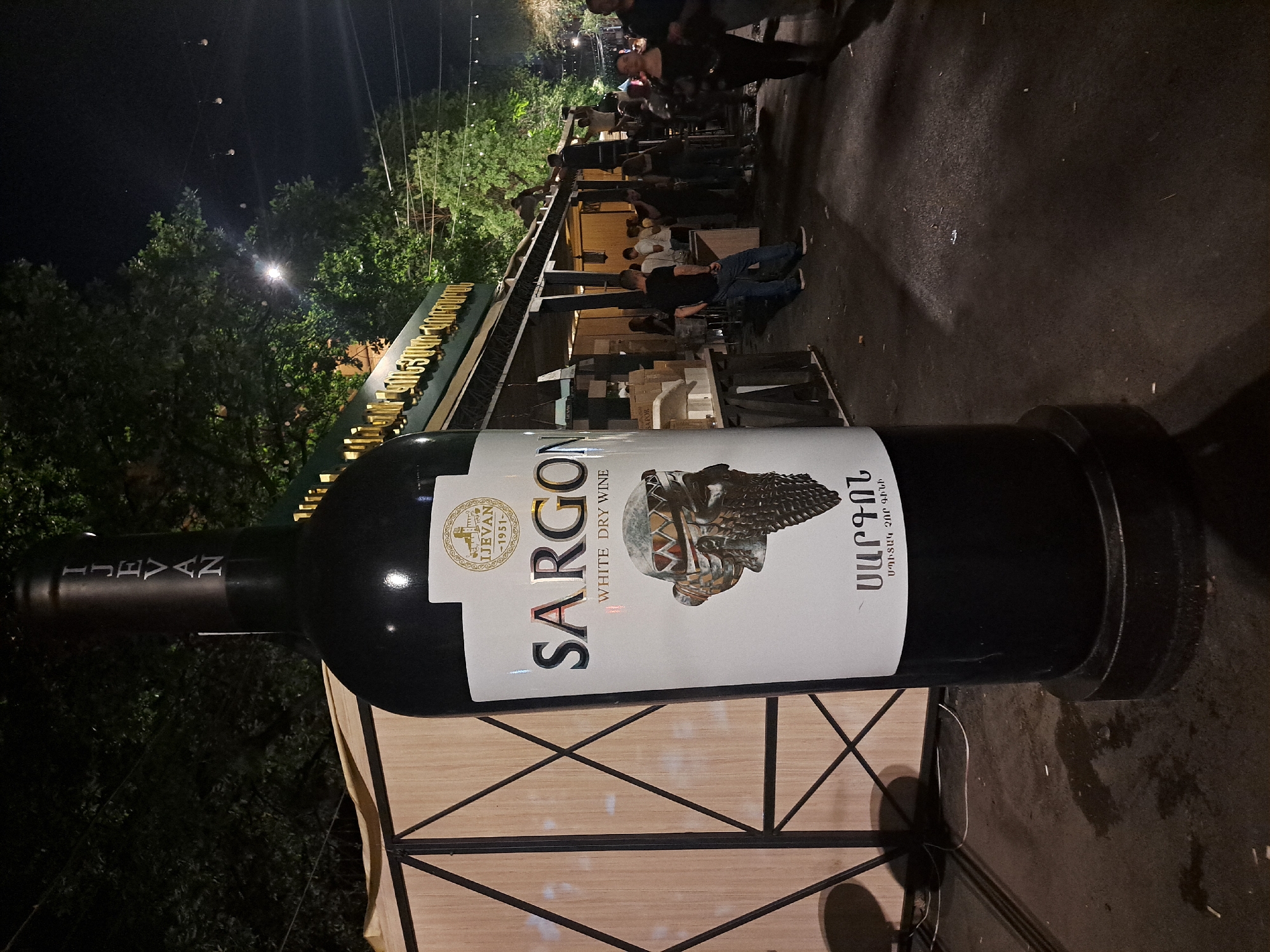 This image captures an outdoor night scene in Yerevan, Armenia, focusing on a large promotional replica of a dark wine bottle. The bottle, positioned centrally in the foreground, features a white label with "IJEVAAN - 1951" and a fortress-like logo at the top. Below this, "SARGON" is displayed in prominent silver-gold lettering, followed by "WHITE DRY WINE" and an illustration of an ancient, textured helmet. The label's base includes "ՍԱՐԳՈՆ" and "ՍՊԻՏԱԿ ՉՈՐ ԳԻՆԻ" in Armenian. The bottle's neck also bears "IJEVAAN" written vertically.
To the left, a light-colored wooden structure with dark, angular cross-bracing is partially visible. In the midground, a green-fronted building or temporary stall extends horizontally, displaying "ԻՋԵՎԱՆ ԳԻՆՈՒ" (Ijevan Wine) in golden Armenian text on its fascia, identifying the brand.
Numerous individuals are present. Several men are on the roof structure of the green-fronted building, some standing on ladders, engaged in activities such as setup or dismantling. Below, other people, some blurred by motion, are gathered in front of the building and in the open space to the right. One man in a black t-shirt and blue jeans stands with his back to the camera in the mid-left.
The ground is dark asphalt or pavement, showing scattered small debris and irregular damp patches. The environment is illuminated by overhead streetlights and string lights, with dense green trees visible against the dark night sky. The setting strongly suggests a street festival or market, likely during an event like "Yerevan Wine Days," which commonly takes place on Saryan Street or similar public areas in Yerevan.