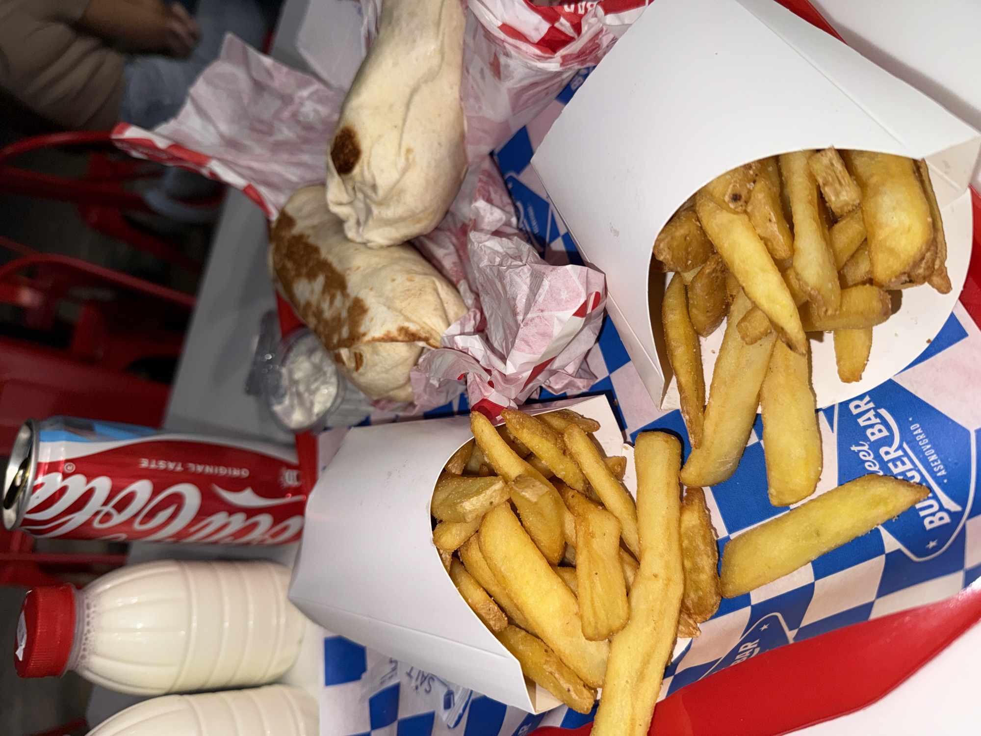 The image displays a fast-food meal, likely consumed indoors. The primary subjects are various food and drink items arranged on a table or tray.
In the foreground, there are two white cardboard boxes filled with golden-brown, thick-cut French fries. Next to these, two wrapped food items, resembling burritos or large wraps, are partially unwrapped, showing browning on their tortillas and resting on crumpled red and white checkered paper.
Beverages include a red Coca-Cola can, clearly labeled "Coca-Cola" and "ORIGINAL TASTE." Below the can are two white, ribbed plastic bottles with red caps, containing a milky white liquid, possibly milk or a milkshake.
The food and drinks are situated on a red surface, which is largely covered by blue and white checkered paper. This paper features text, including "BURGER BAR" and "Asenovgrad," identifying the likely origin of the meal. A person's arm, dressed in a beige sleeve, is partially visible in the blurry background on the left side of the image, suggesting someone is present at the meal. The lighting appears to be typical indoor artificial light. The scene captures a casual dining event, possibly at a burger restaurant or enjoying takeout.