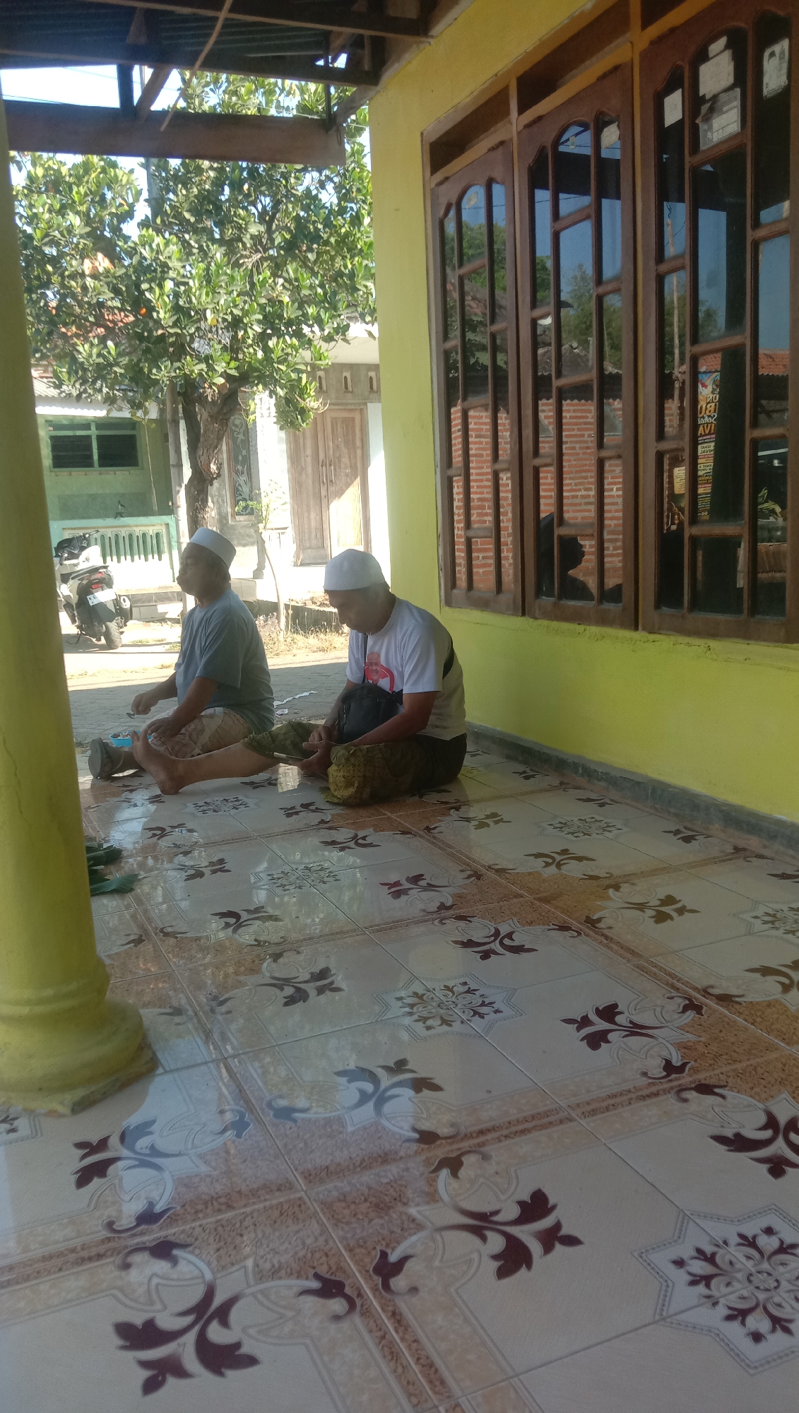 Two men, wearing white skullcaps, are sitting on the tiled floor of a porch in Ngempit, Indonesia. The man on the left wears a gray t-shirt and patterned shorts, while the man on the right wears a white t-shirt with a graphic design and dark pants. The man on the right is holding a mobile phone and appears to be looking at it. A dark bag is positioned next to him. Both men have their legs crossed. The porch has a yellow wall and a wooden window with multiple panes. Trees and a motorbike are visible in the background. The floor is decorated with a repeating pattern of floral motifs in brown and cream colors. The overall scene suggests a casual, outdoor setting on a sunny day.
