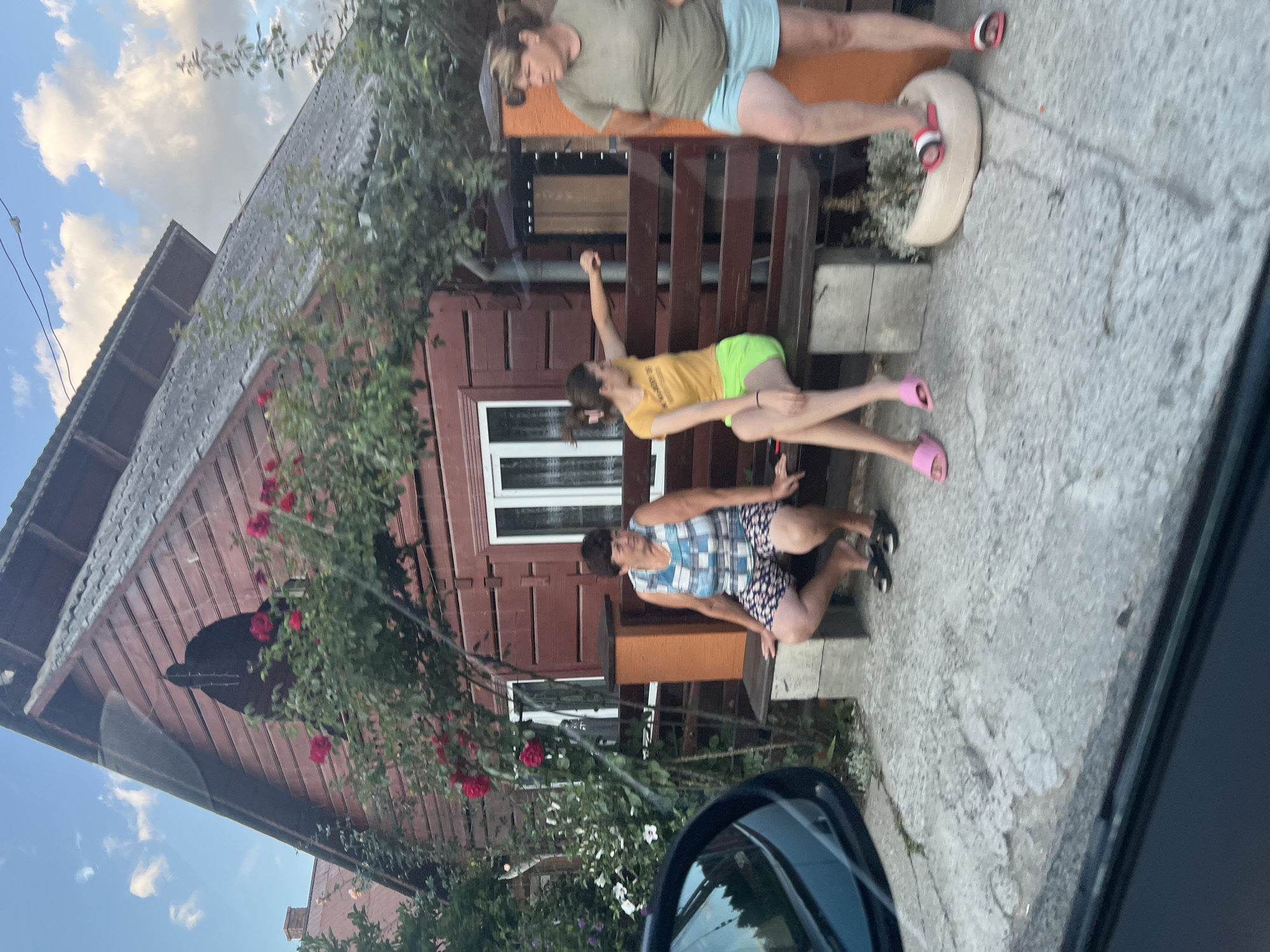In Rona de Sus, Romania, three women are visible on a sunny day, gathered outside a traditional wooden house. The house has a prominent, steeply pitched roof covered with dark shingles and features several windows. Red roses climb the facade of the house and over a trellis. Two of the women are seated on a wooden bench, while the third stands to the right of the bench. The woman on the far left of the bench wears a blue and white checkered tank top and patterned shorts. Next to her, a younger woman in a yellow t-shirt and bright green shorts is also seated. The standing woman on the right wears a plain olive-green t-shirt and light blue shorts. The ground in front of the bench is paved with concrete. A circular planter with a small plant is located to the right of the standing woman. The scene appears to be captured from a vehicle, as a portion of a car's side mirror is visible in the lower left corner of the image.