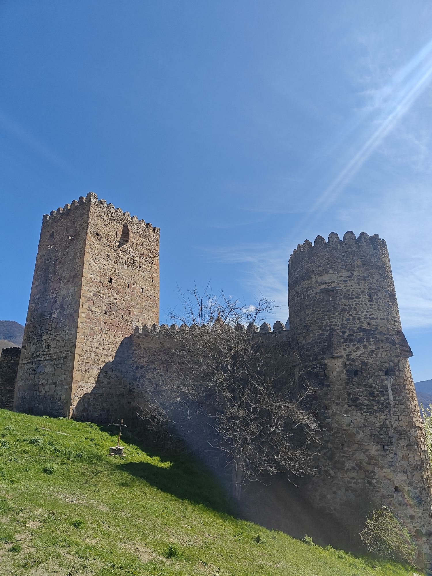 A low-angle shot depicts two stone towers connected by a wall with crenellations, situated on a grassy hillside. The tower on the left is rectangular in shape, constructed from reddish-brown stone, and has a small window near the top. The tower on the right is cylindrical, built from gray stone, and also features crenellations. A bare tree stands in front of the wall connecting the towers. A small stone cross stands on the hill in front of the wall. The sky is a clear blue with a few wispy clouds. The scene is set in the city of Unknown, Unknown.