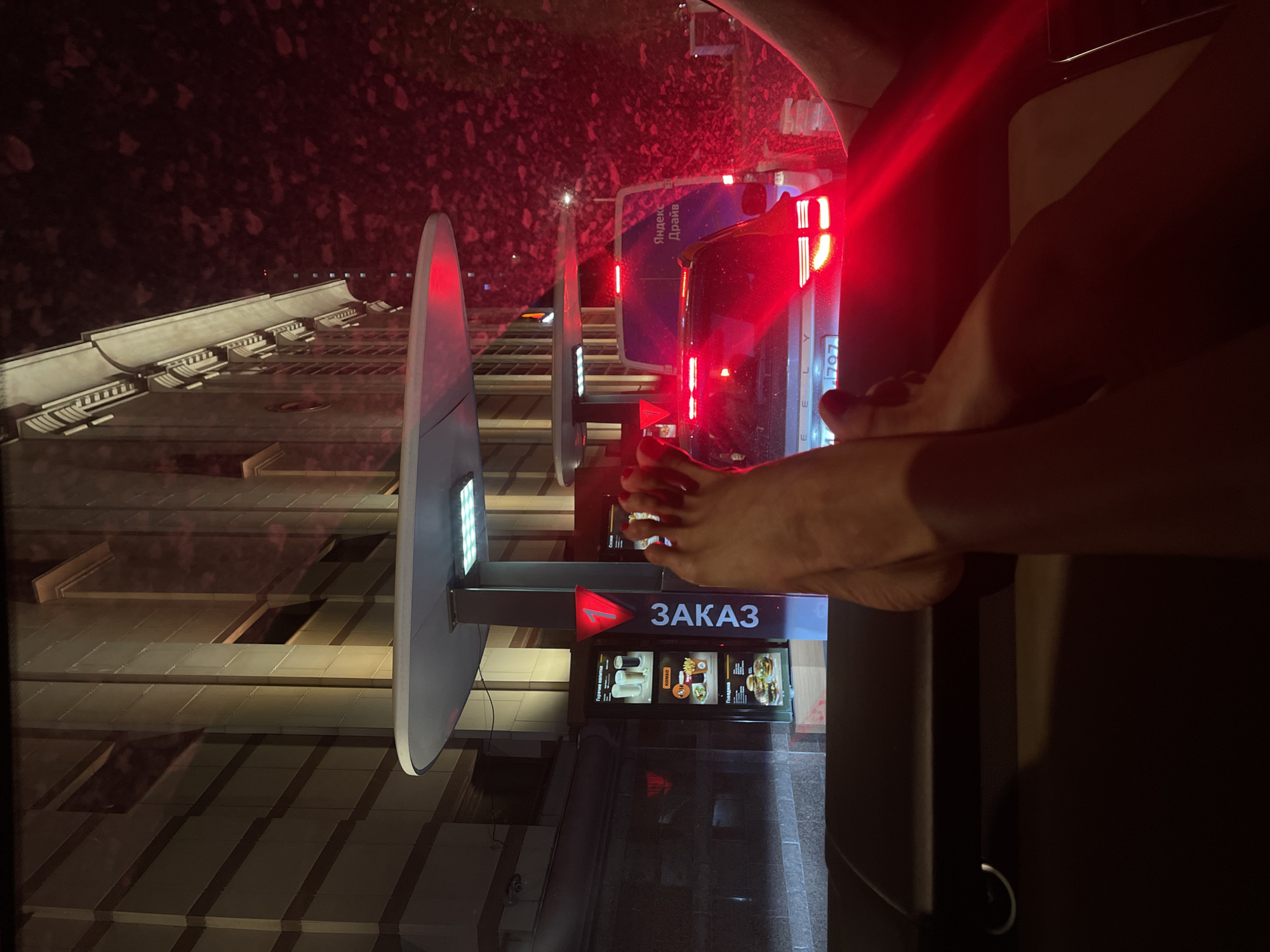 Here is a detailed description of the image:
**Scene Overview:**
The image appears to be taken from inside a car at night, possibly in a drive-through setting. The primary focus is on an outdoor ordering station, with a red light indicating "ЗAКАЗ" (order in Russian). The image also features a building's exterior on the left side of the frame.
**Objects:**
* **Ordering Station:** An outdoor kiosk with a digital menu displaying food items and the word "ЗAКАЗ" (Order) in Russian. A light on top of the sign.
* **Vehicle:** The image is taken from within a car. Parts of the dashboard and possibly the side window are visible. The person inside the car has their feet on the dashboard
* **Building:** A multi-story building with a repetitive window pattern.
* **Vehicle:** A passenger vehicle is behind the car, featuring an illuminated advertisement for Yandex Drive and license plate 707.
**People:**
* **Person in the Car:** Bare feet are visible on the dashboard of the car.
**Environment:**
* **Setting:** A city environment at night.
* **Weather:** Appears to be nighttime.
**Location:**
The presence of Russian text ("ЗAКАЗ" and "Яндекс Драйв") indicates the location is in Russia, and the vehicle that is behind the vehicle with the feet, has license plate 707, indicating it is Moscow, Russia.
