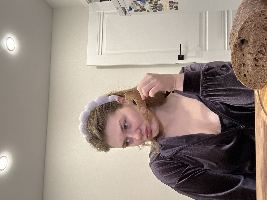 A woman in a purple velvet top and a gray headband holds up a thin slice of bread to her face. The bread is brown with holes. To the right, a loaf of the same bread sits on a wooden surface. A white cabinet with black hardware is in the background. Two lights are visible on the wall.
